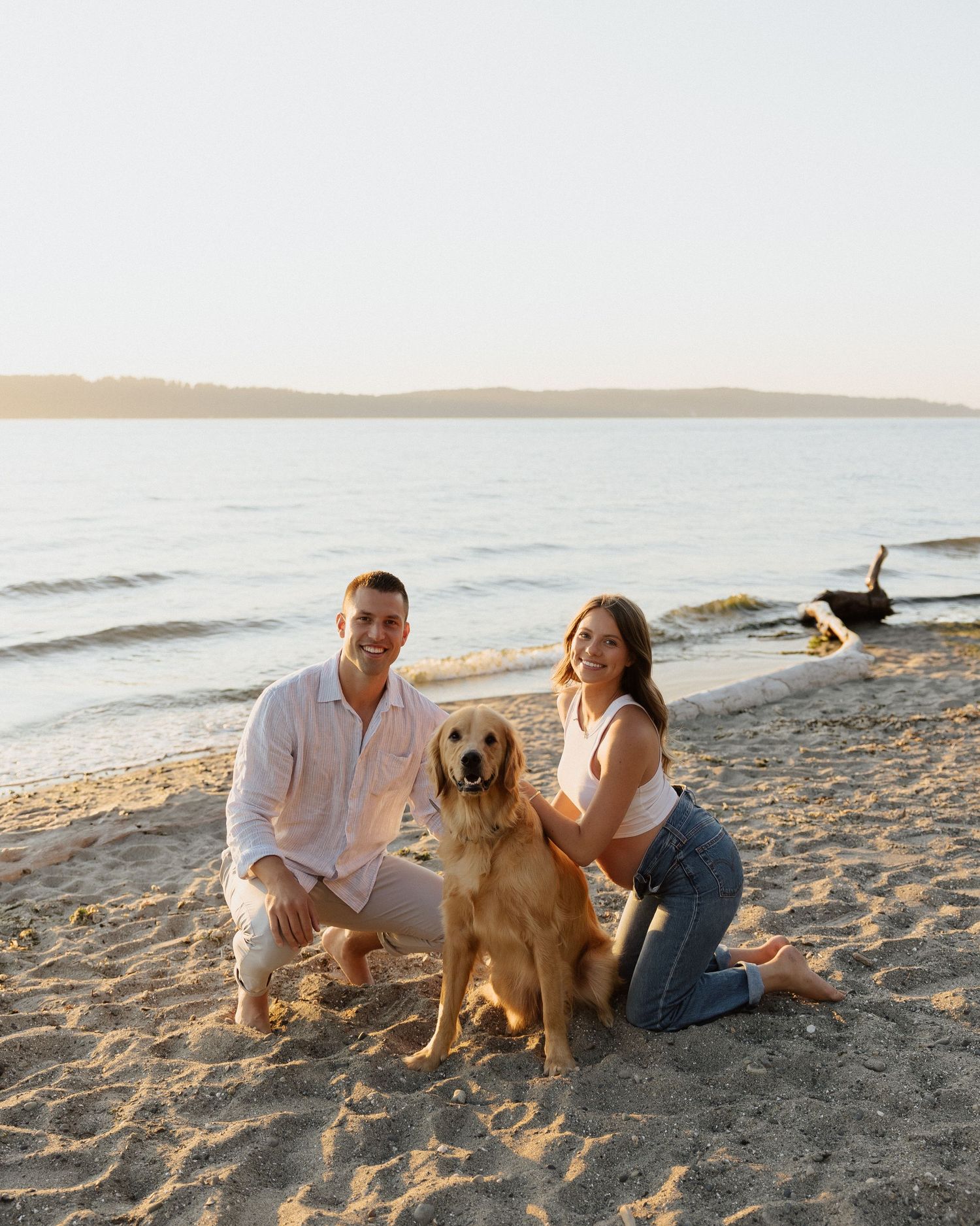 Couple poses with their golden retriever on sandy beach during sunset.