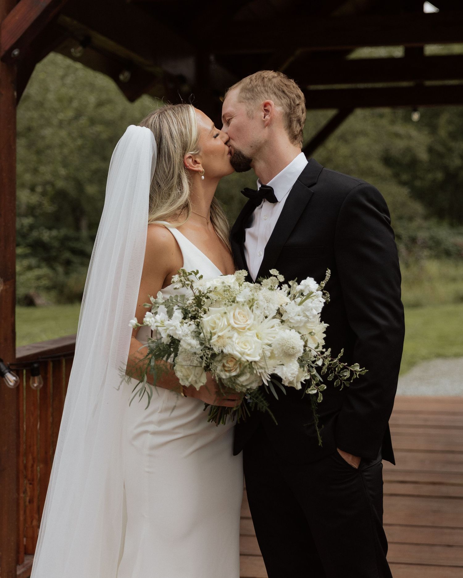 Romantic kiss between newlyweds on wooden bridge with white floral bouquet.