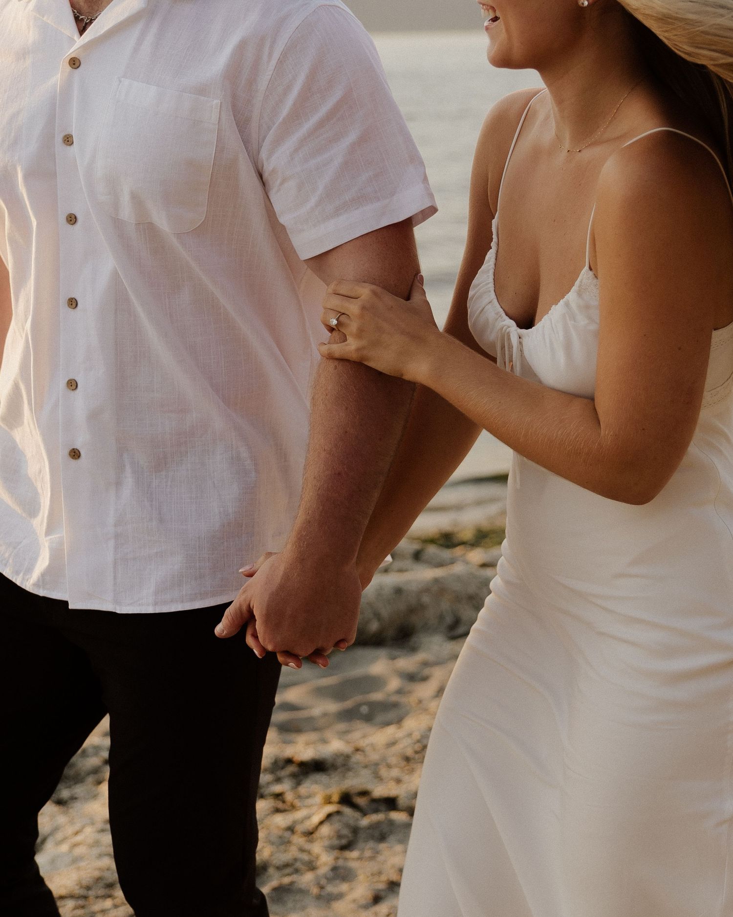 Close-up of a couple holding hands on a beach, wearing coordinating white outfits.
