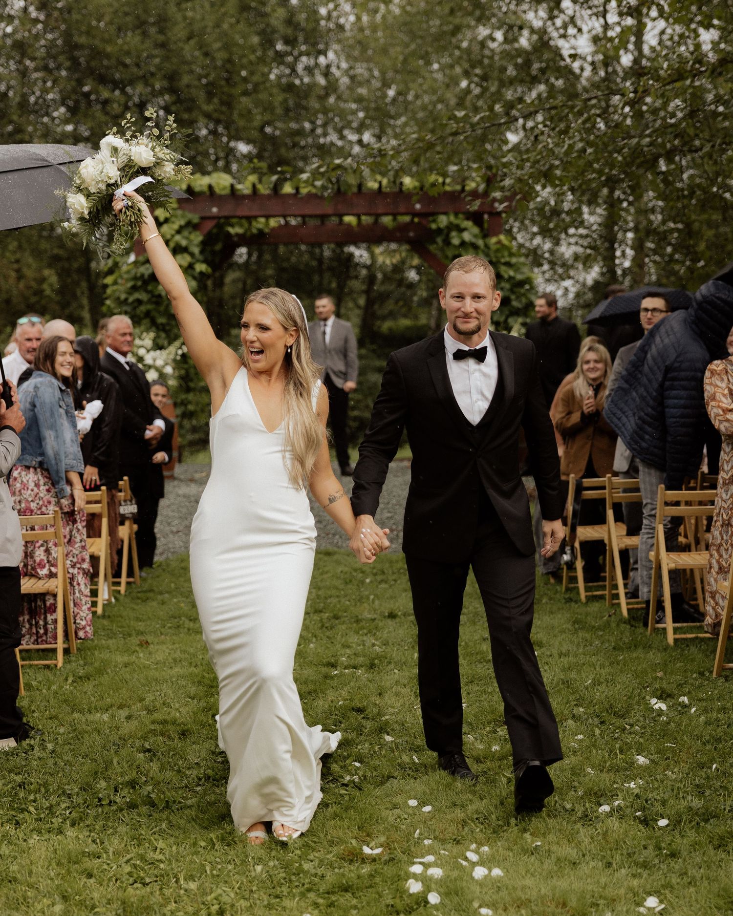 Triumphant couple exits wedding ceremony on flower-strewn grass path.