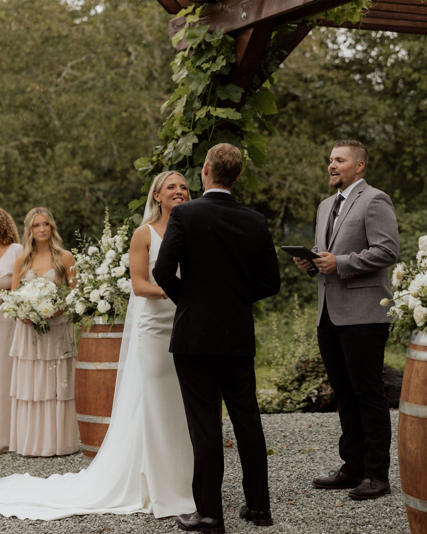 Wedding ceremony under wooden pergola with floral decorations and champagne colored bridesmaid dresses.