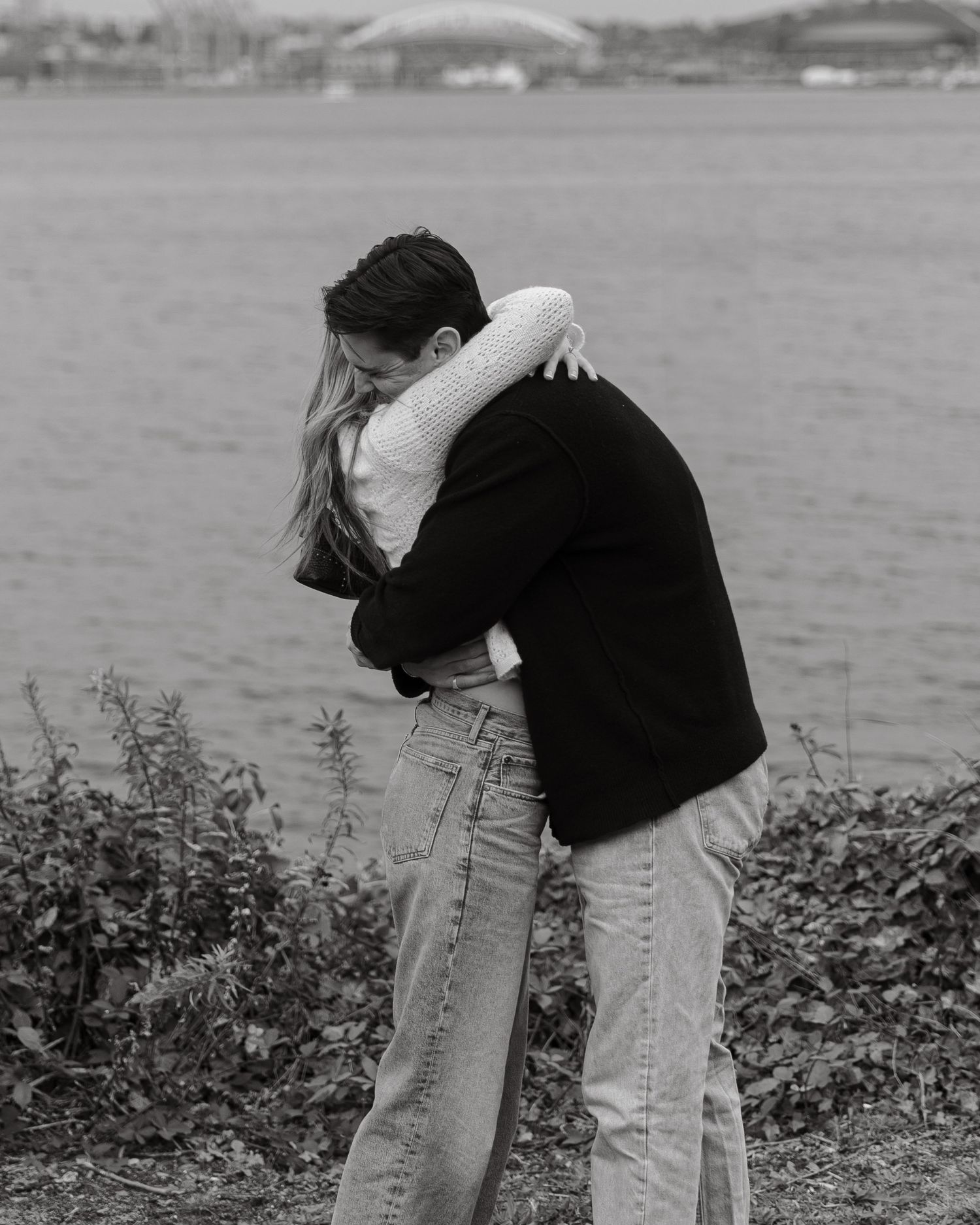 Two people embrace emotionally on a hilltop overlooking a waterfront city vista.