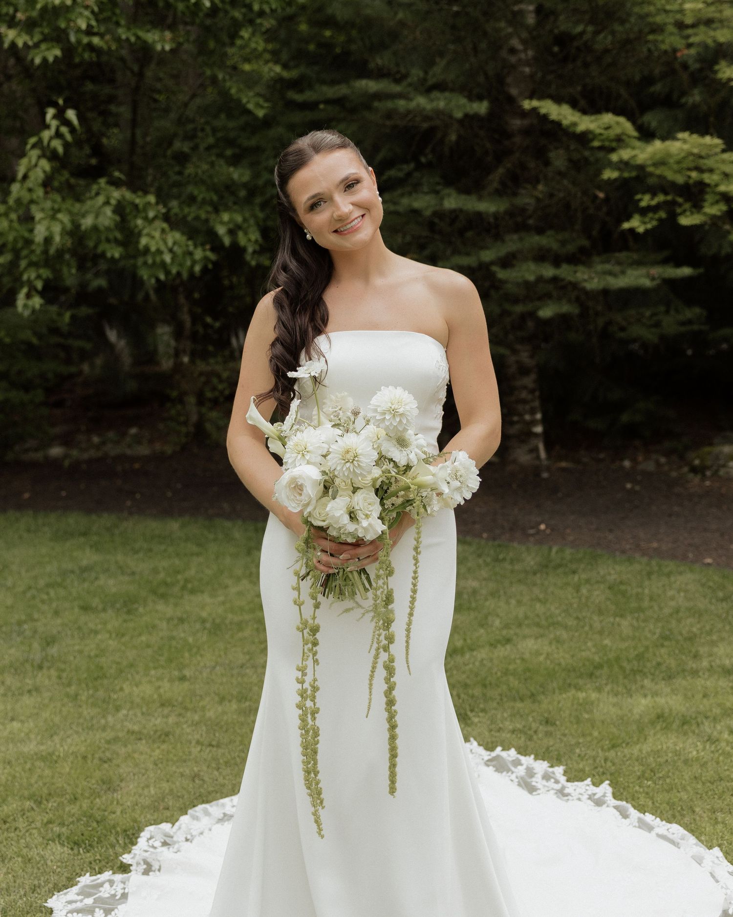 Radiant bride in strapless white mermaid gown holding cascading white bouquet while posing on lush green lawn.