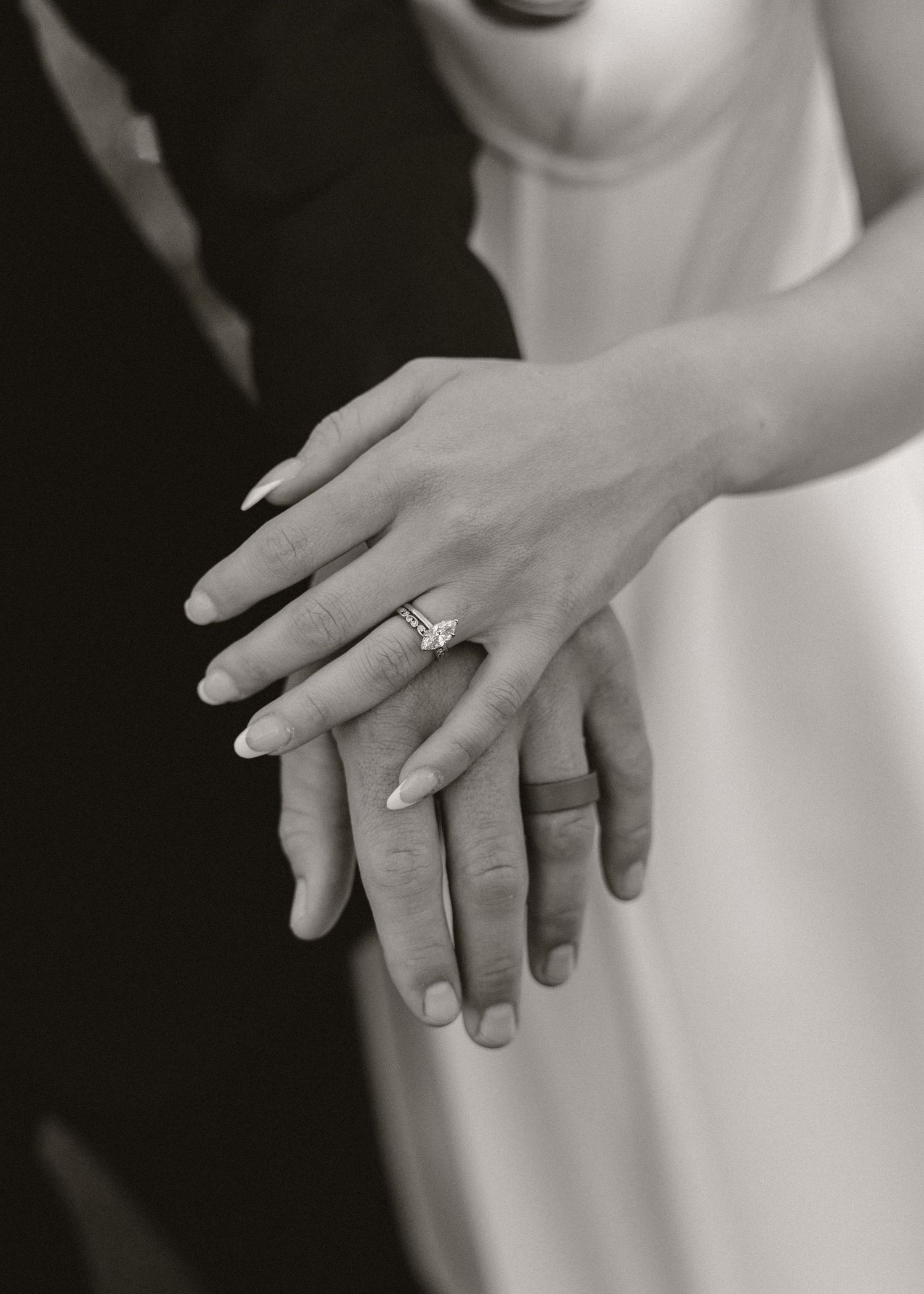 Black and white photo of bride and groom hands together, showcasing a sparkling diamond engagement ring and wedding bands.