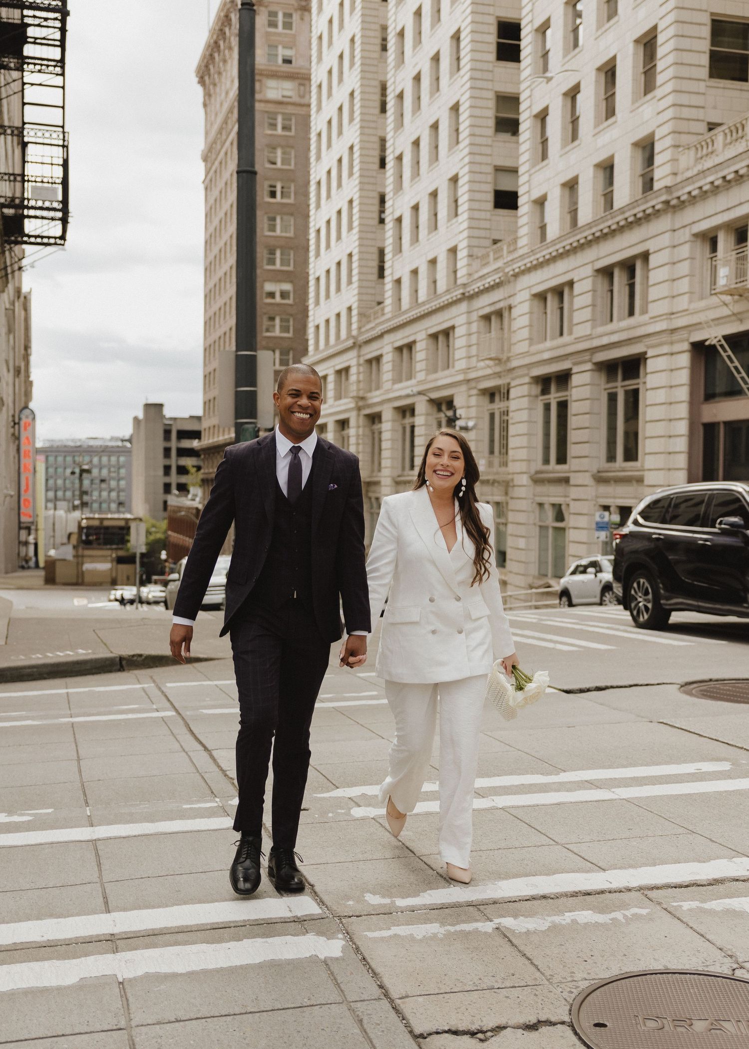 Couple in wedding attire walking hand-in-hand across a crosswalk in an urban city setting on their wedding day.