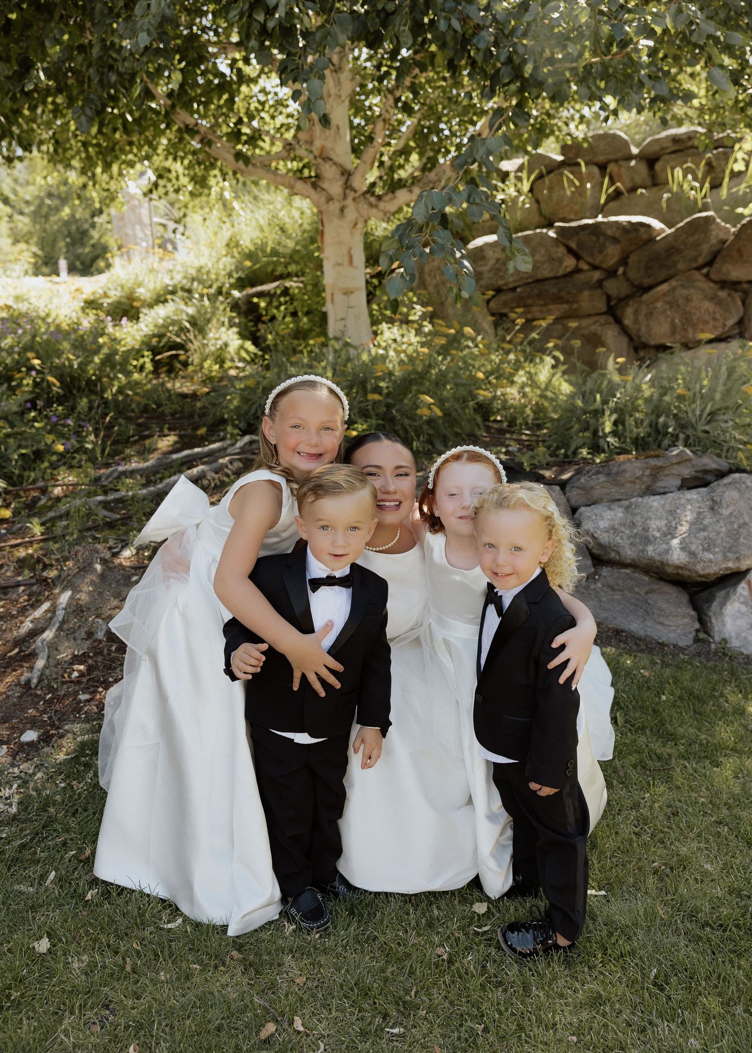 Bride poses with flower girls in white dresses and ring bearers in tuxedos outdoors at a garden wedding.