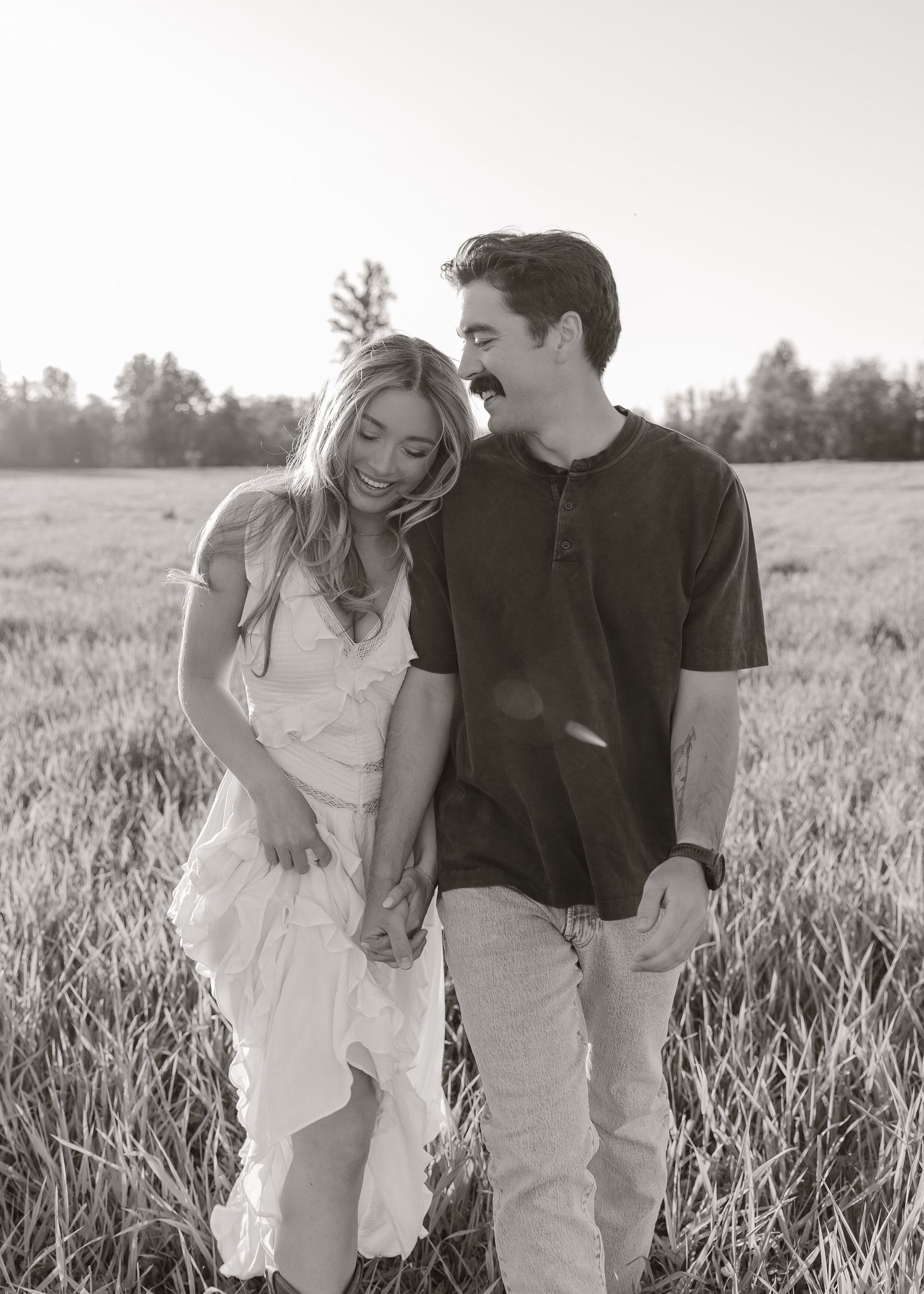 Black and white photo of a smiling couple holding hands walking through a grassy field at sunset.