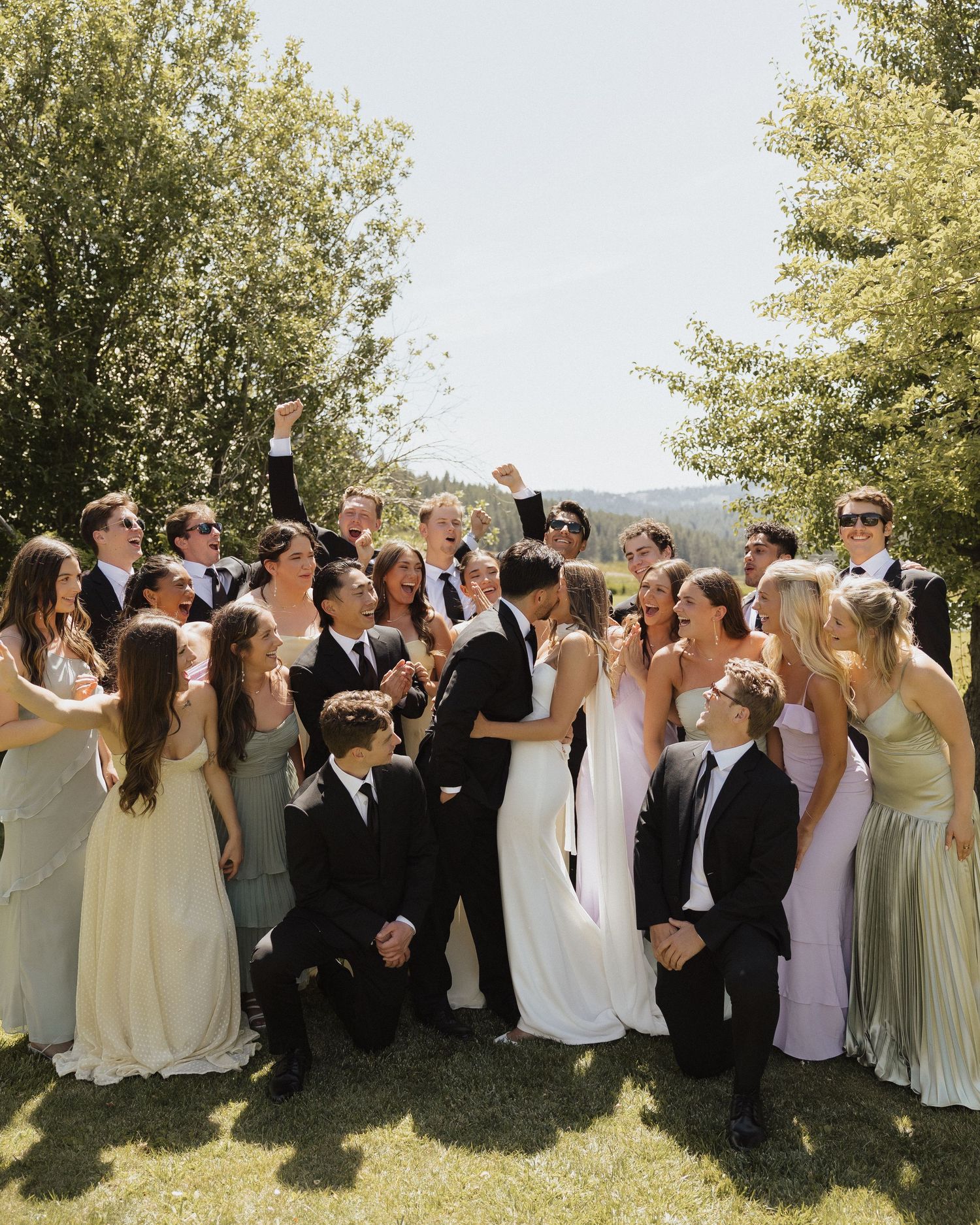 Large wedding party celebrates outdoors with bride and groom kissing surrounded by bridesmaids and groomsmen on sunny day.