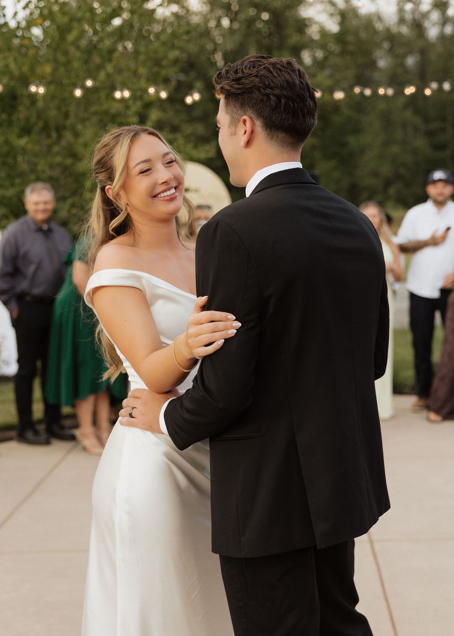 Smiling bride in off-shoulder white gown shares first dance with groom under string lights at outdoor wedding reception.