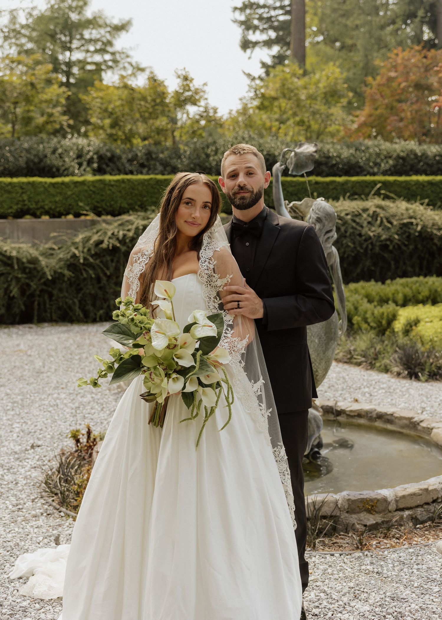 Bride in lace veil and white gown holding green and white bouquet with groom in black suit at garden fountain.