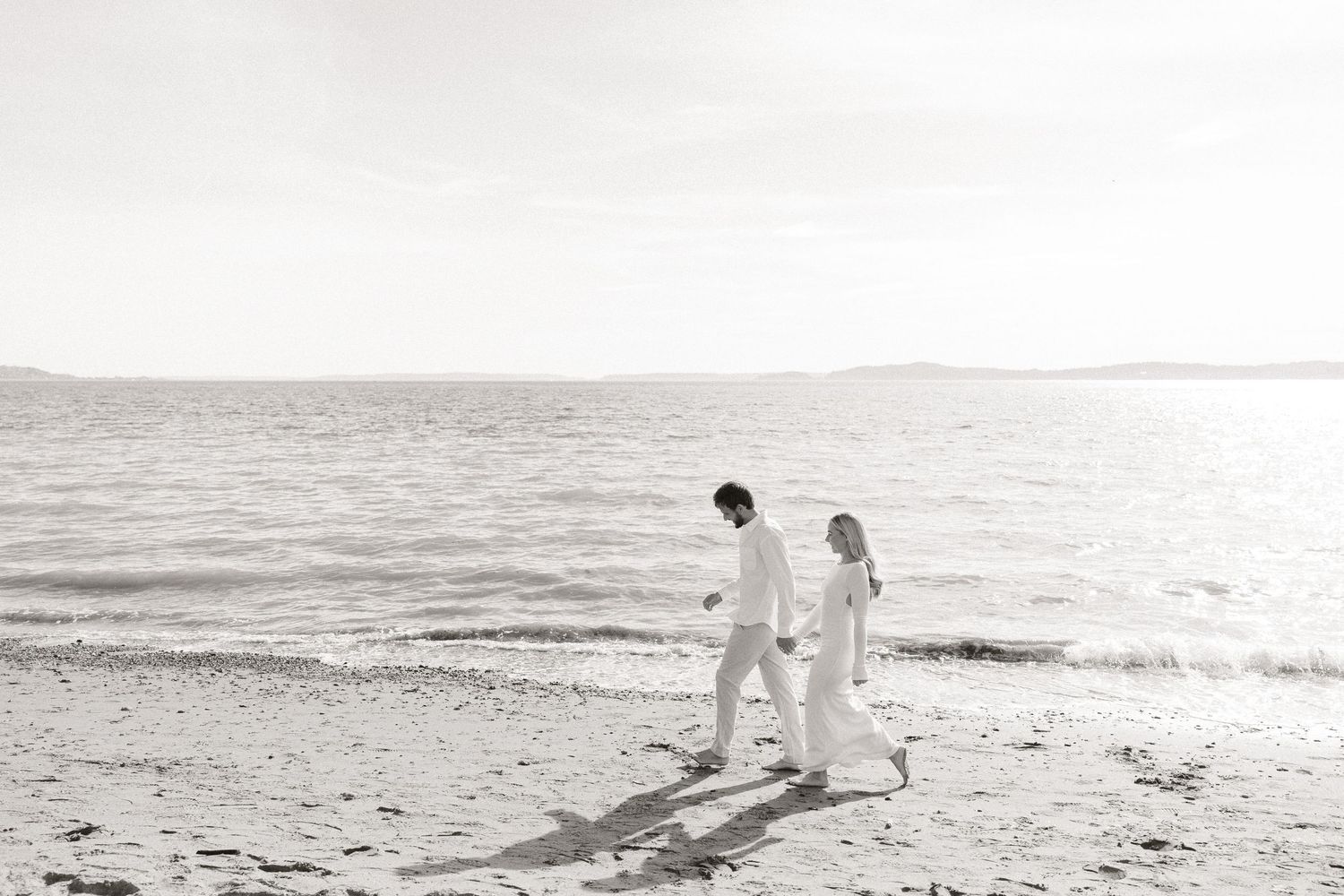 Couple walking hand in hand along a serene beach shoreline in a black and white photo.