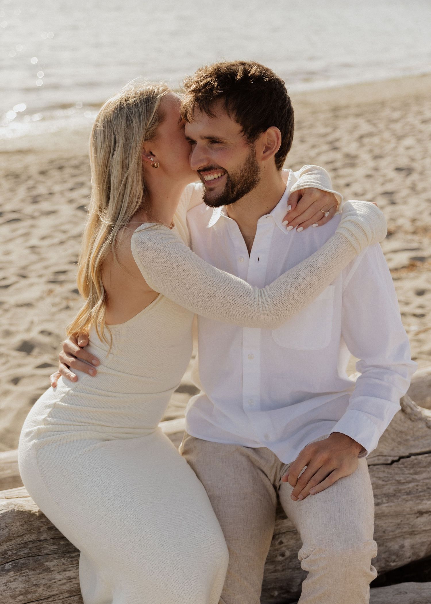 Smiling couple sitting on driftwood at the beach, woman in backless white dress embracing man.