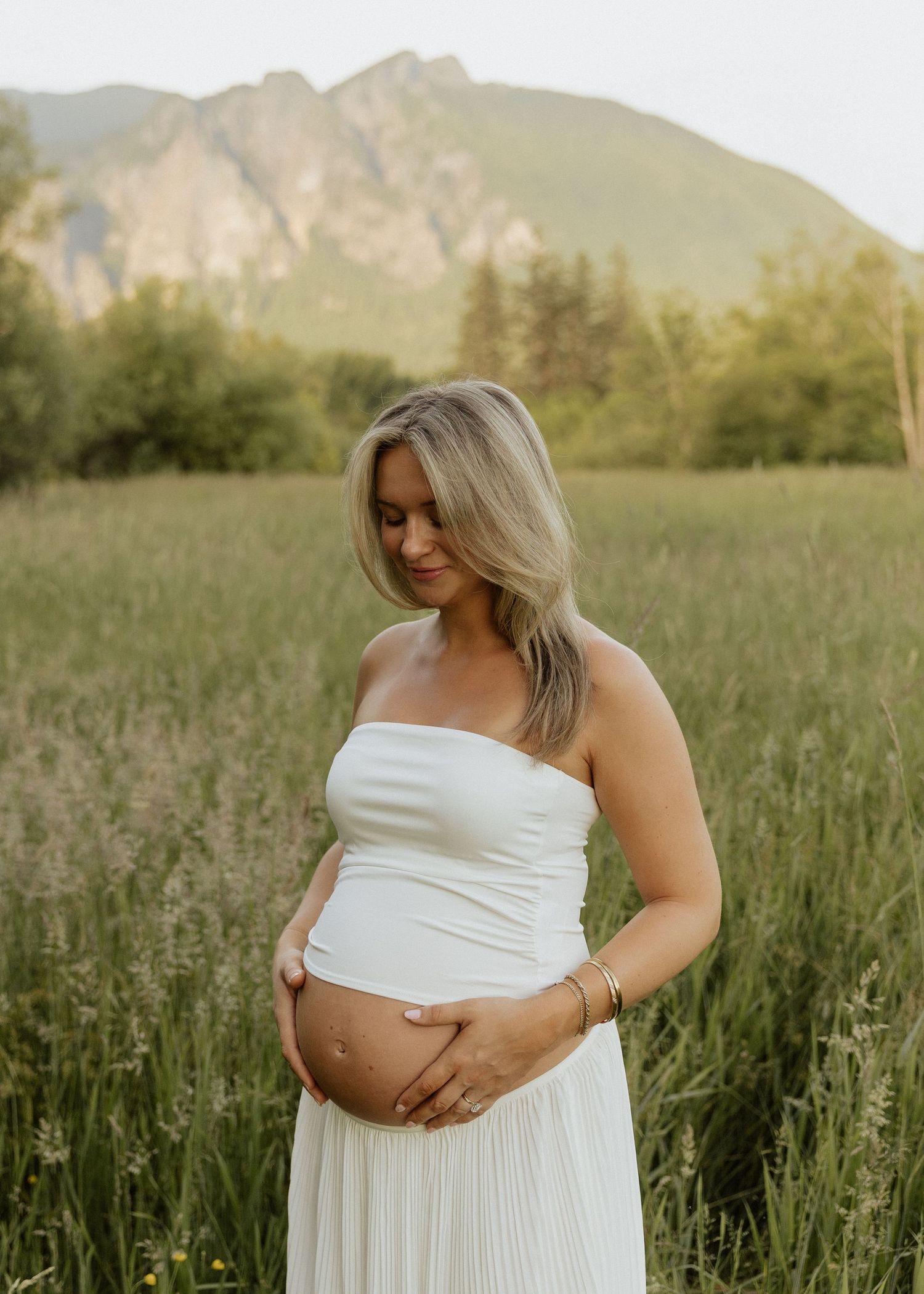 Pregnant woman in white strapless dress cradling baby bump in a meadow with mountain backdrop at sunset.