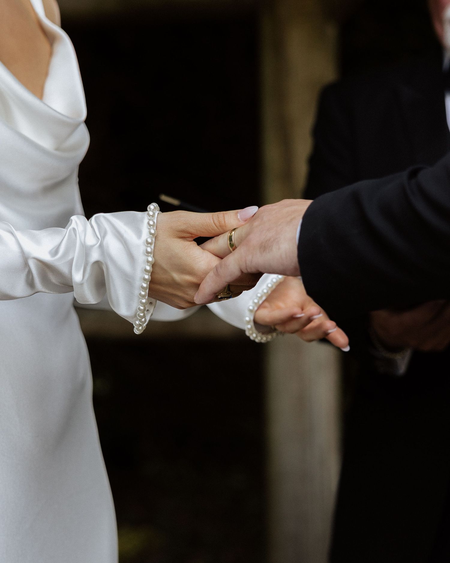 Bride and groom holding hands during wedding ceremony, bride wearing white satin dress and pearl bracelets.