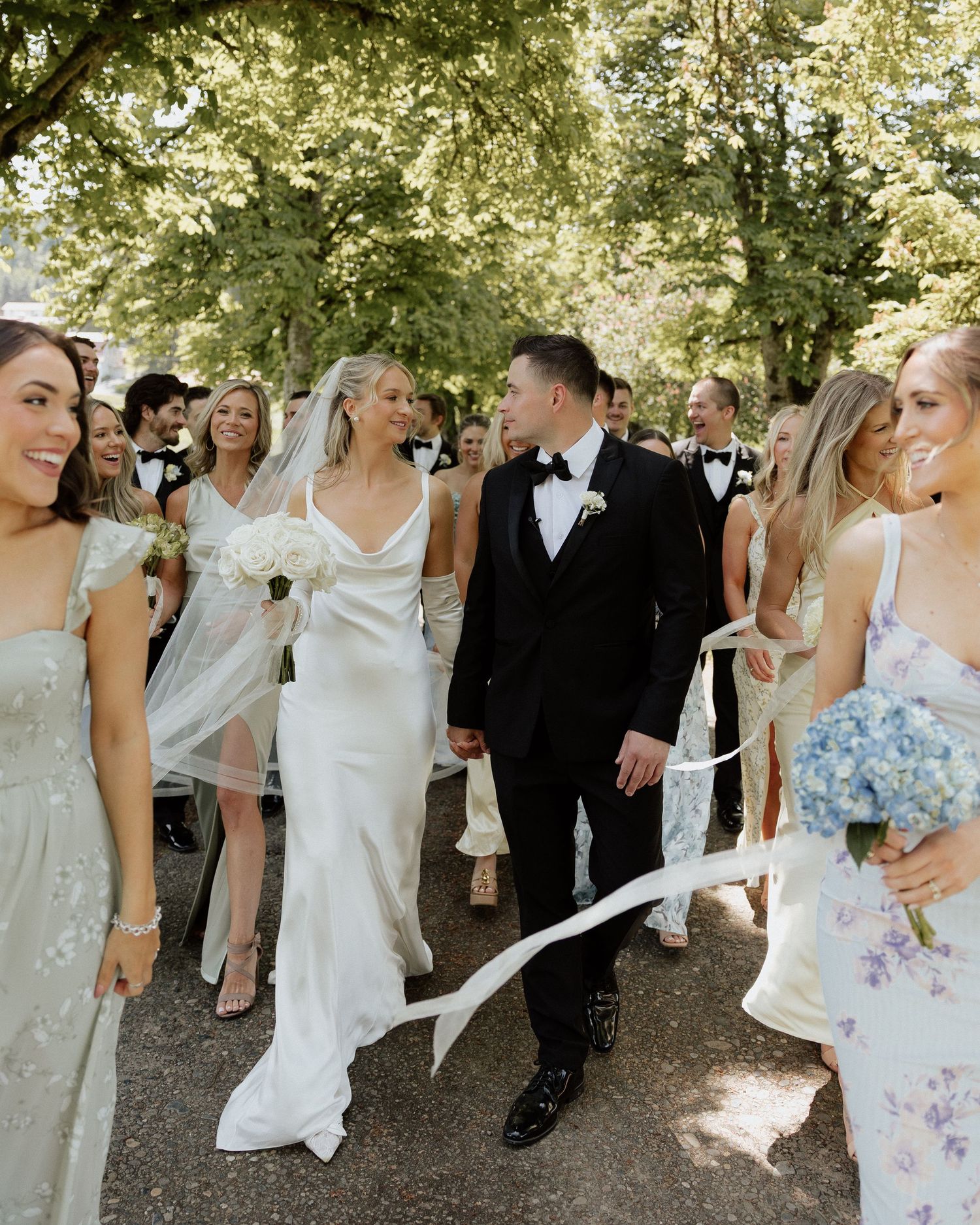 Bride and groom walk hand-in-hand surrounded by wedding party guests outdoors under lush green trees.