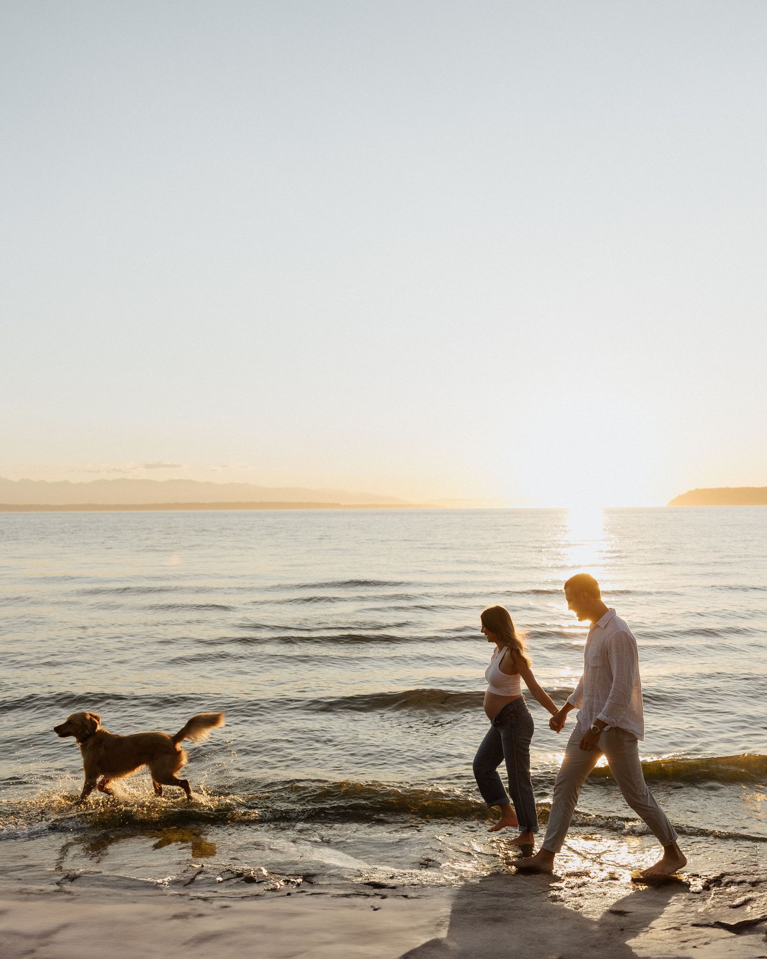 Couple walks along beach at sunset with their dog splashing in the waves.