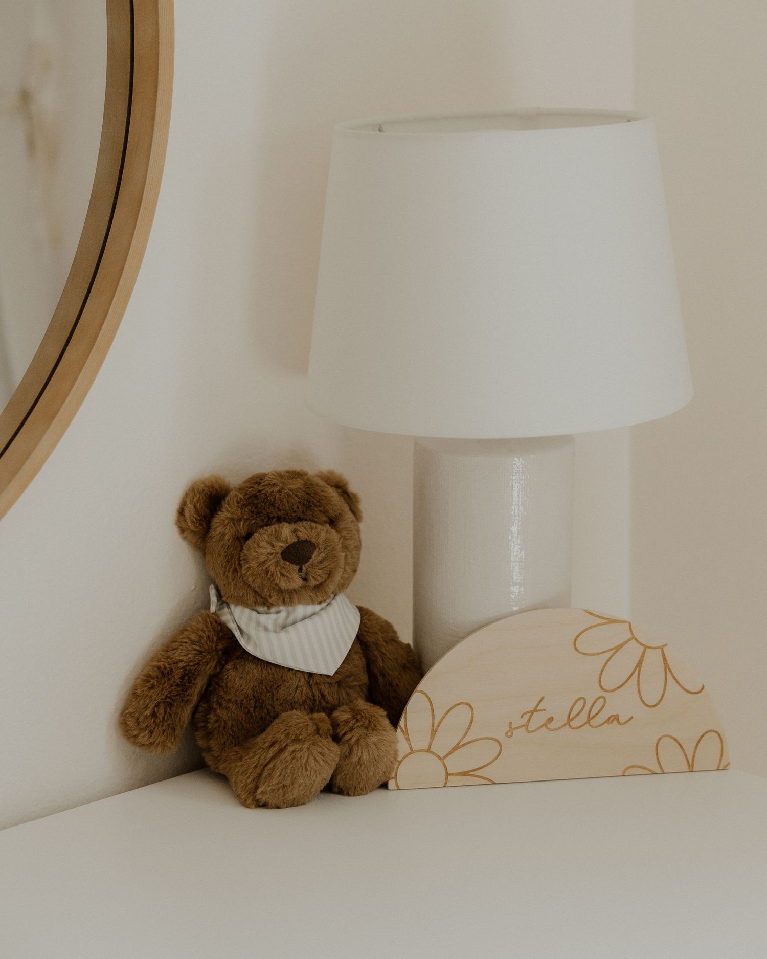 Brown teddy bear with bandana sits next to white lamp on nursery shelf.