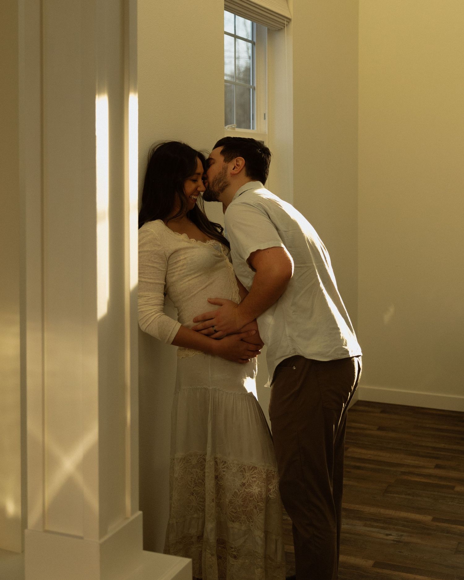 A tender maternity moment captured in warm sunlight streaming through windows.