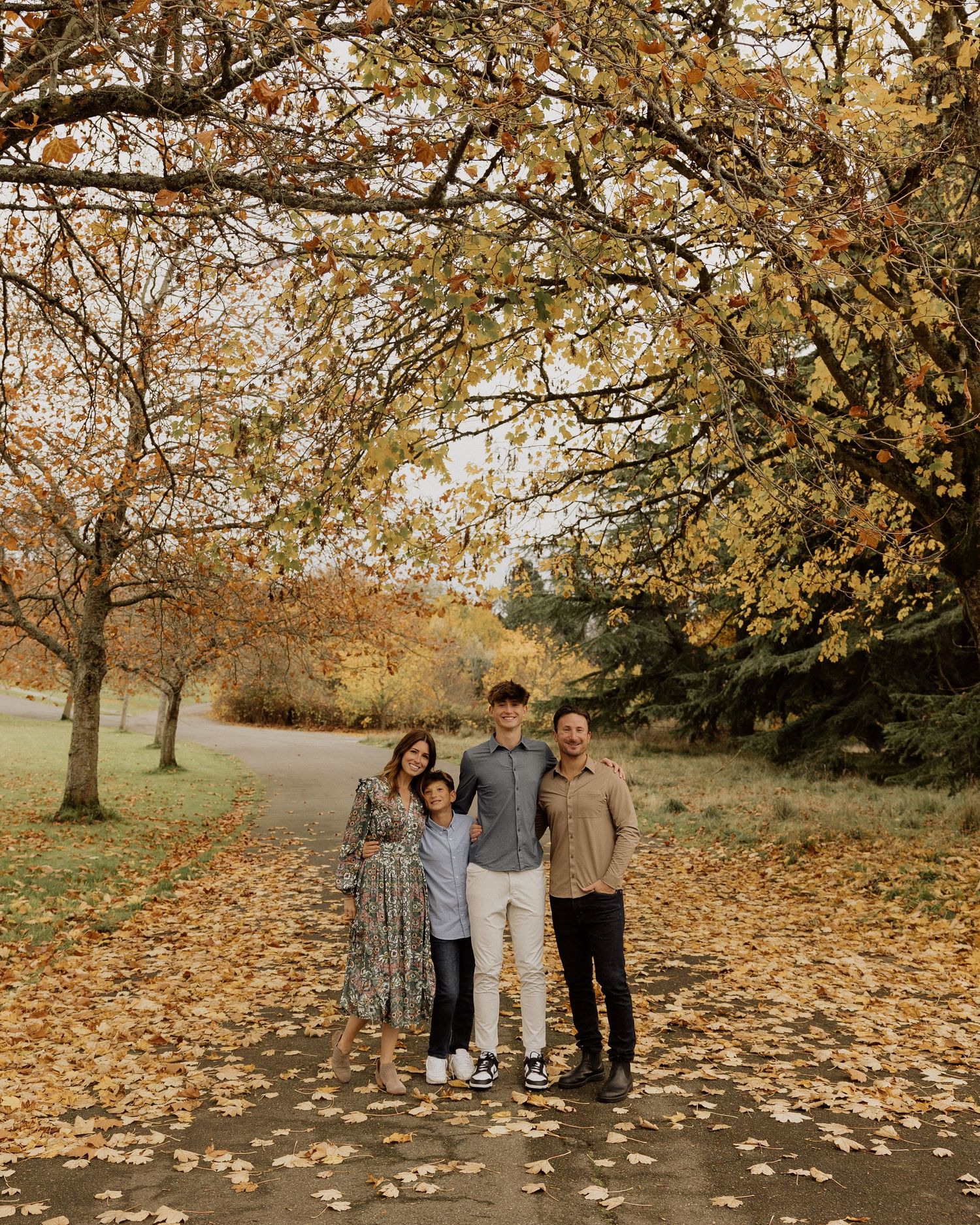 Family autumn portrait beneath golden trees with fallen leaves on ground.