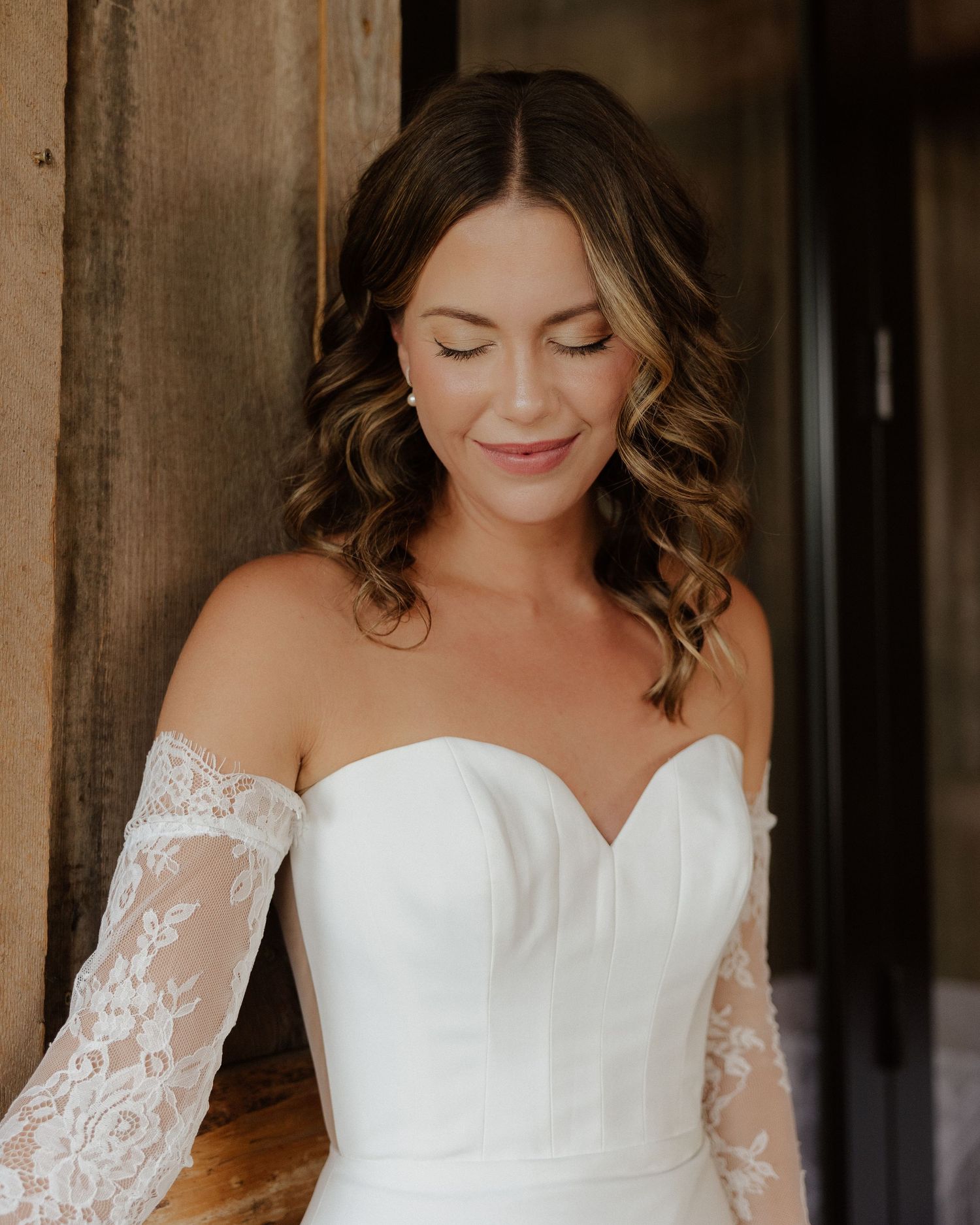 Close-up portrait of bride in off-shoulder lace sleeve wedding dress with natural makeup and curled hair.