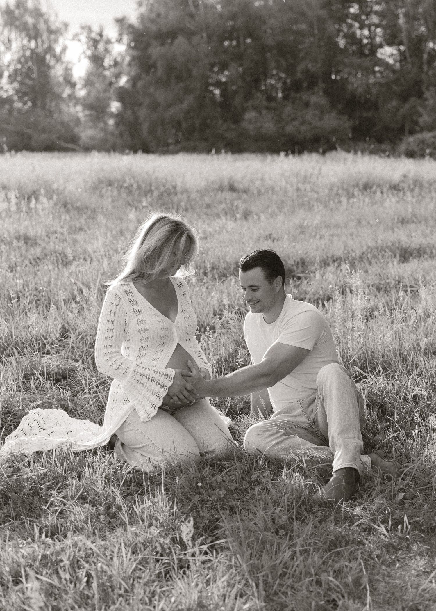 Black and white maternity photo of couple sitting in a field, man tenderly touching pregnant woman's belly.