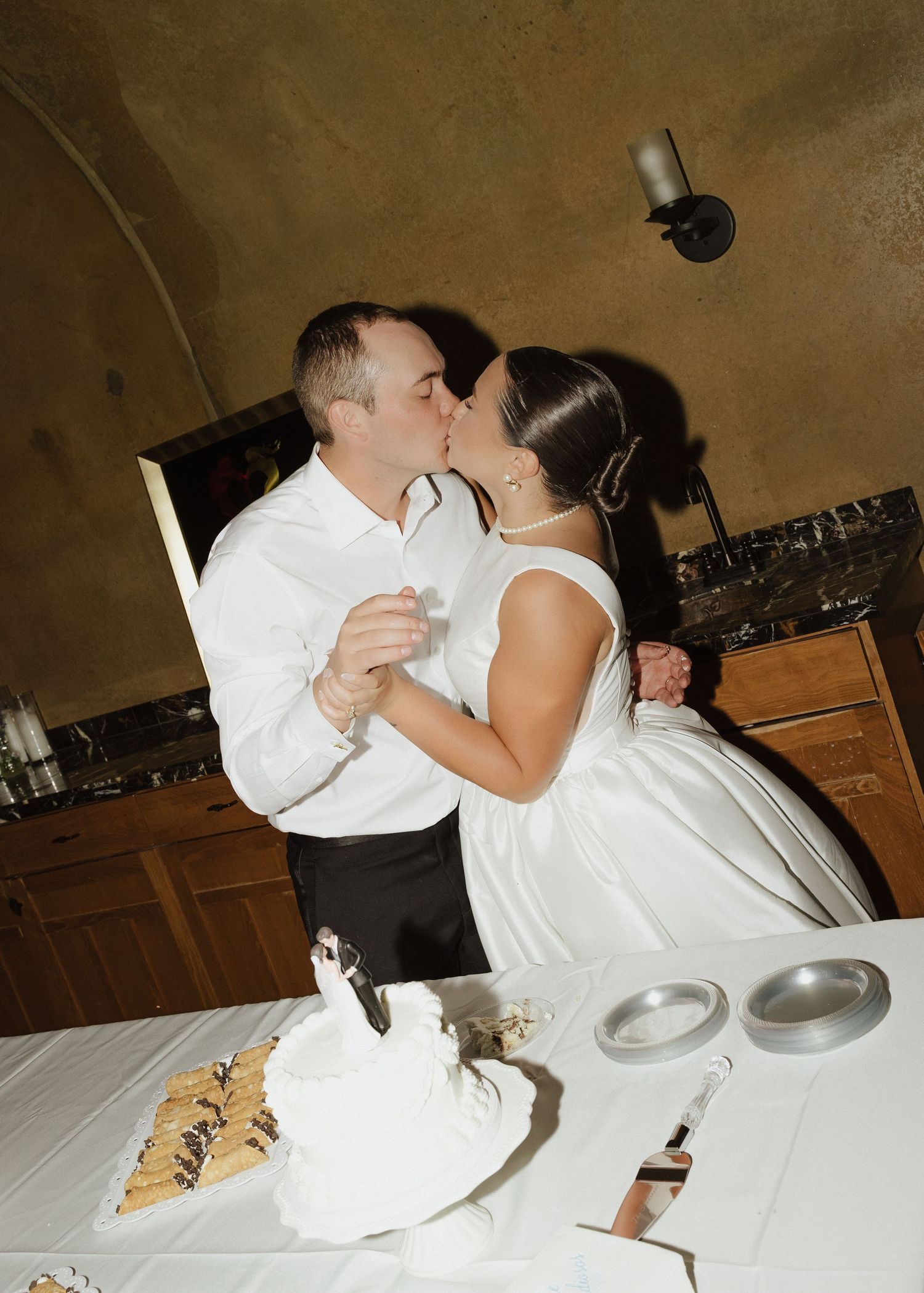 Newlyweds share a romantic kiss while cutting their wedding cake at an indoor reception venue.