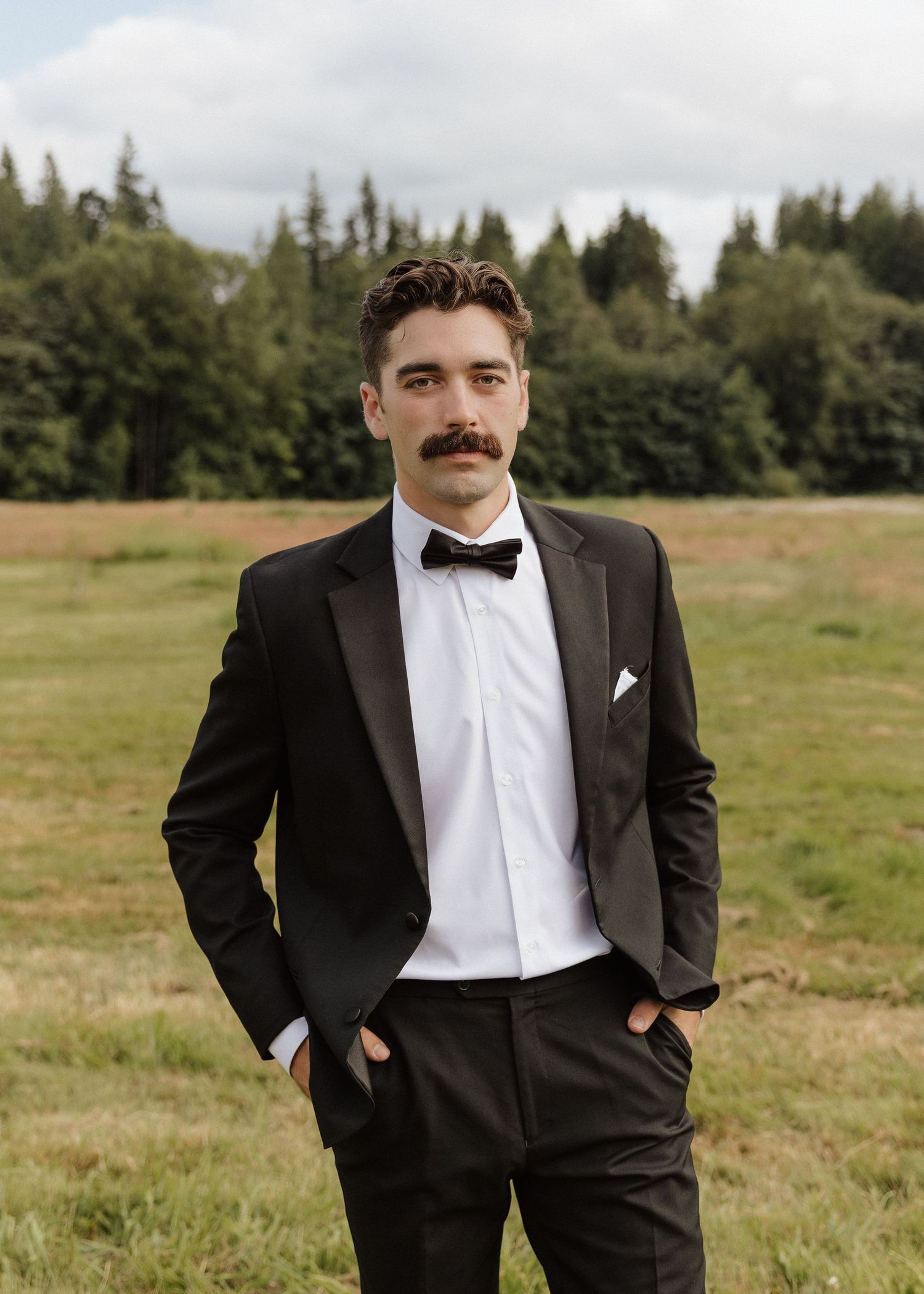 Groom in black tuxedo with bow tie and white pocket square standing in a green field.