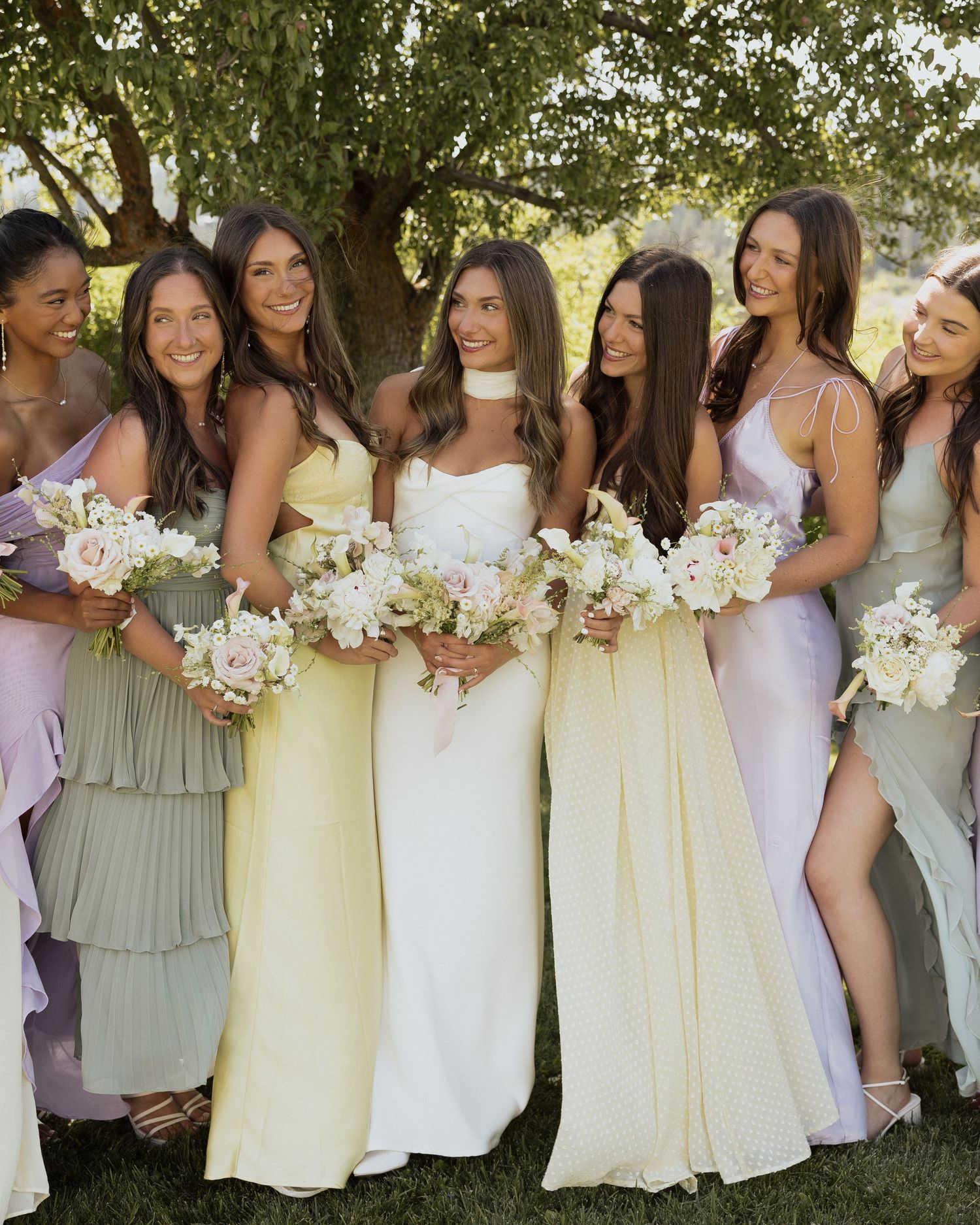 Bride in white gown poses with six bridesmaids in pastel yellow, green, and lavender dresses holding floral bouquets outdoors.