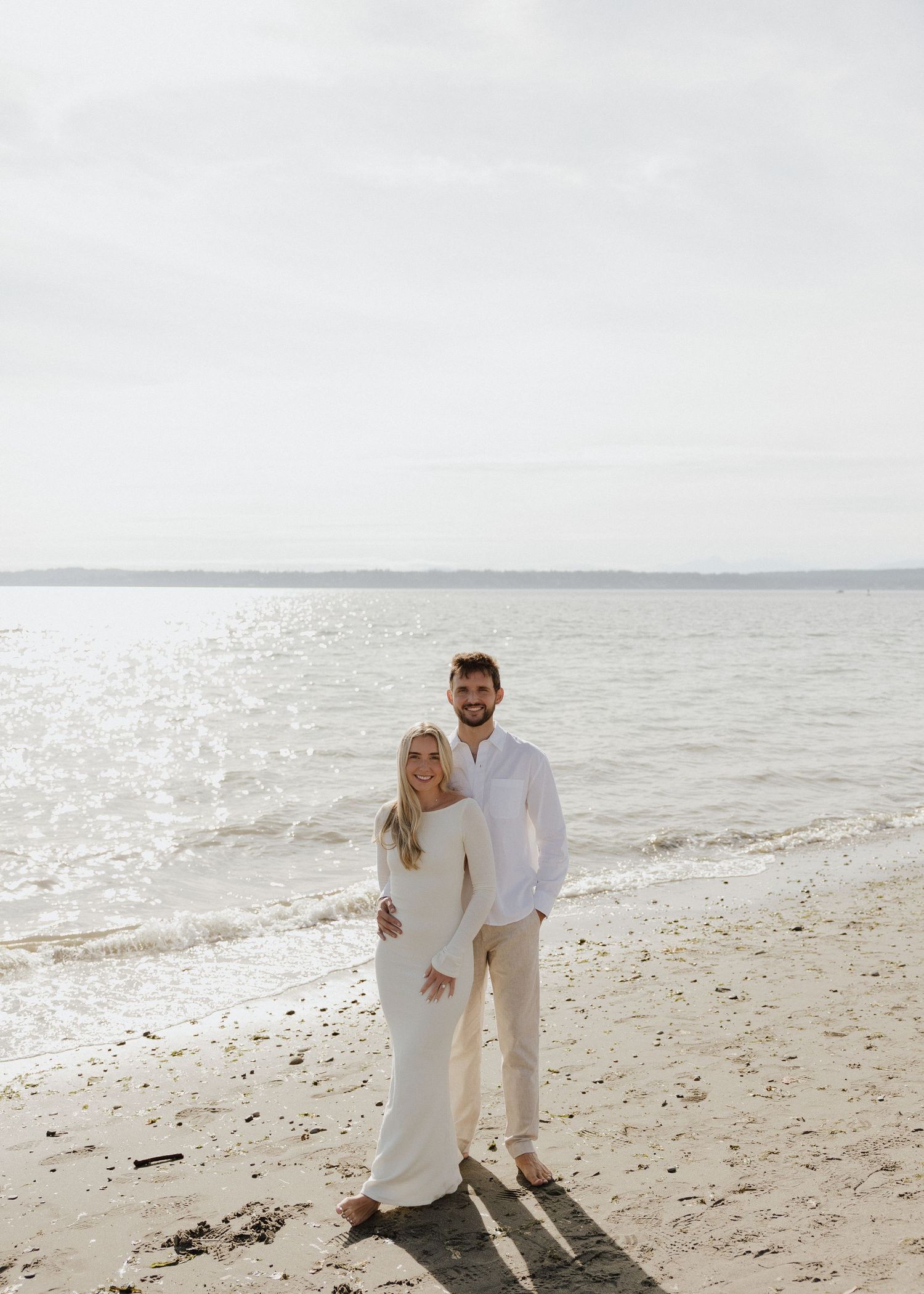 Smiling couple posing together on a sandy beach, dressed in coordinating white and beige outfits, calm ocean behind them.