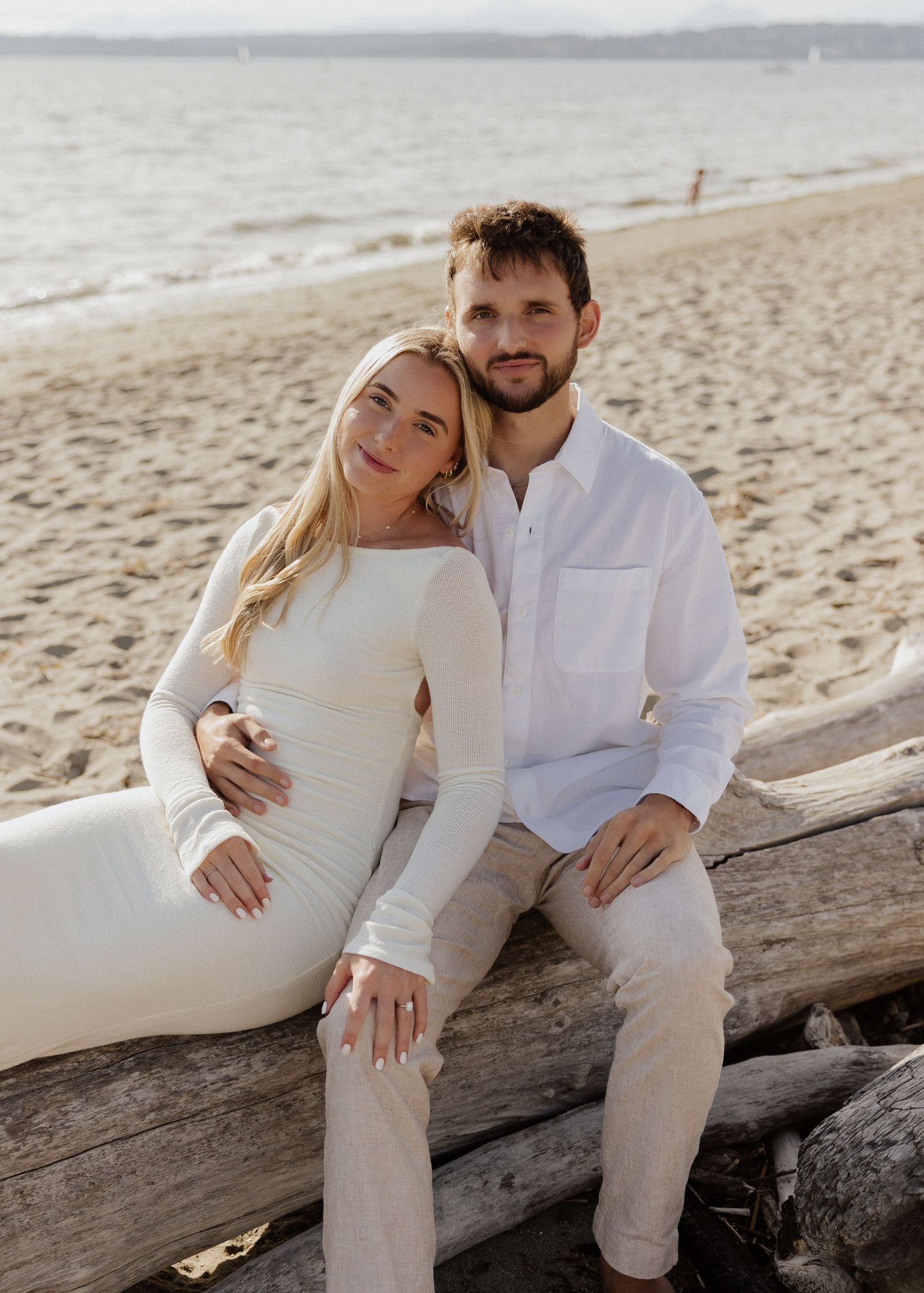 Pregnant woman in white dress and man sitting on driftwood log at a sandy beach.