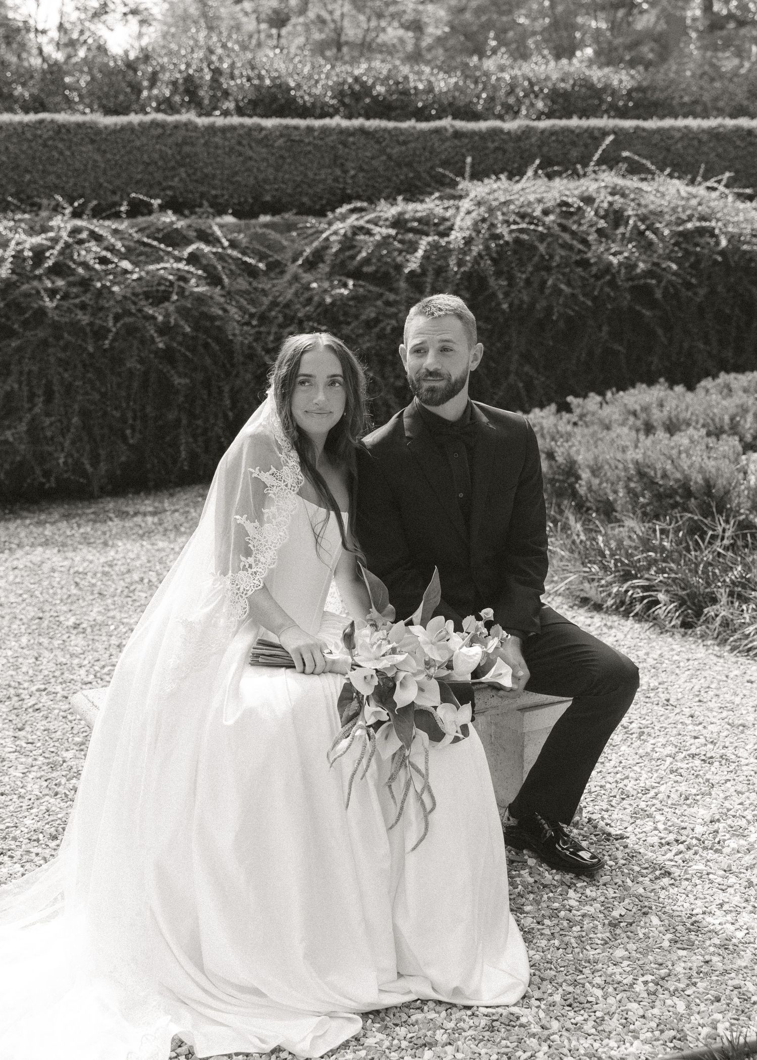 Black and white photo of bride and groom seated in formal garden with hedges, bride holding bouquet.