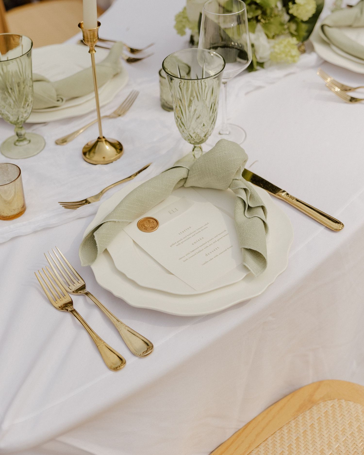 Elegant wedding table setting with gold cutlery, sage green napkin, wax seal menu card, and crystal glassware.