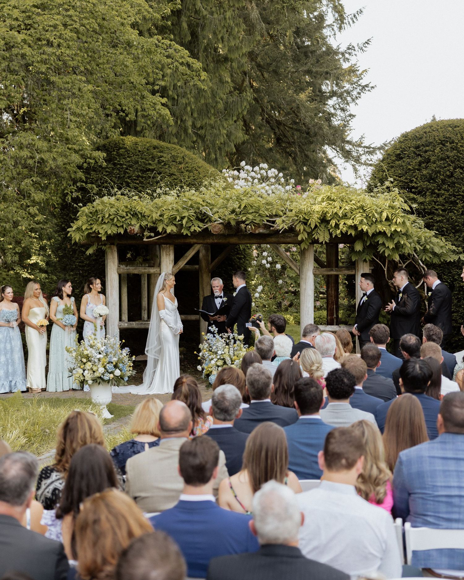 Outdoor wedding ceremony with guests seated on lawn, bridal party in blue, couple at floral altar under vine-covered pergola.