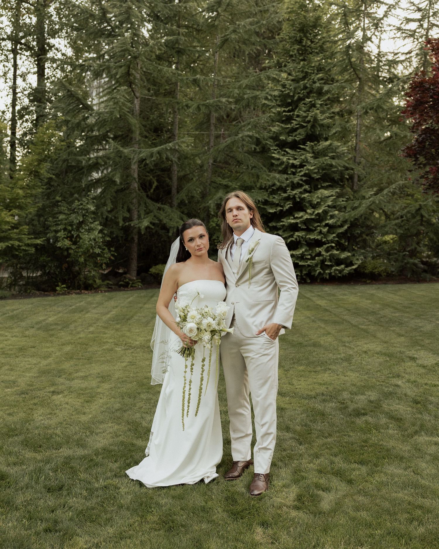 Bride and groom pose on green lawn surrounded by tall evergreen trees.