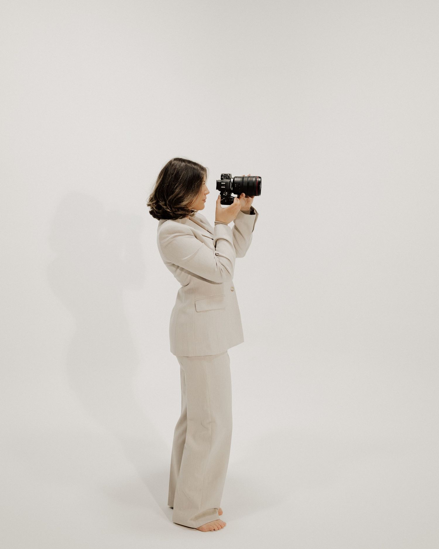 Photographer in cream colored suit holding camera against white backdrop.