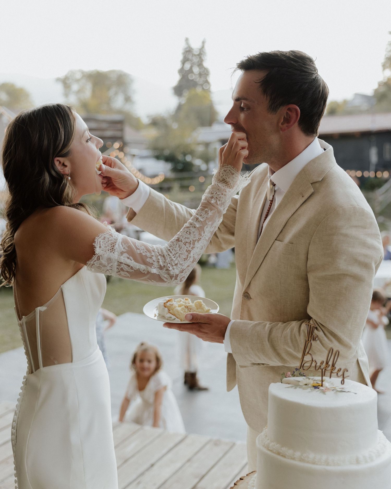 Newlyweds playfully share wedding cake during reception on outdoor deck with white wedding cake in foreground.