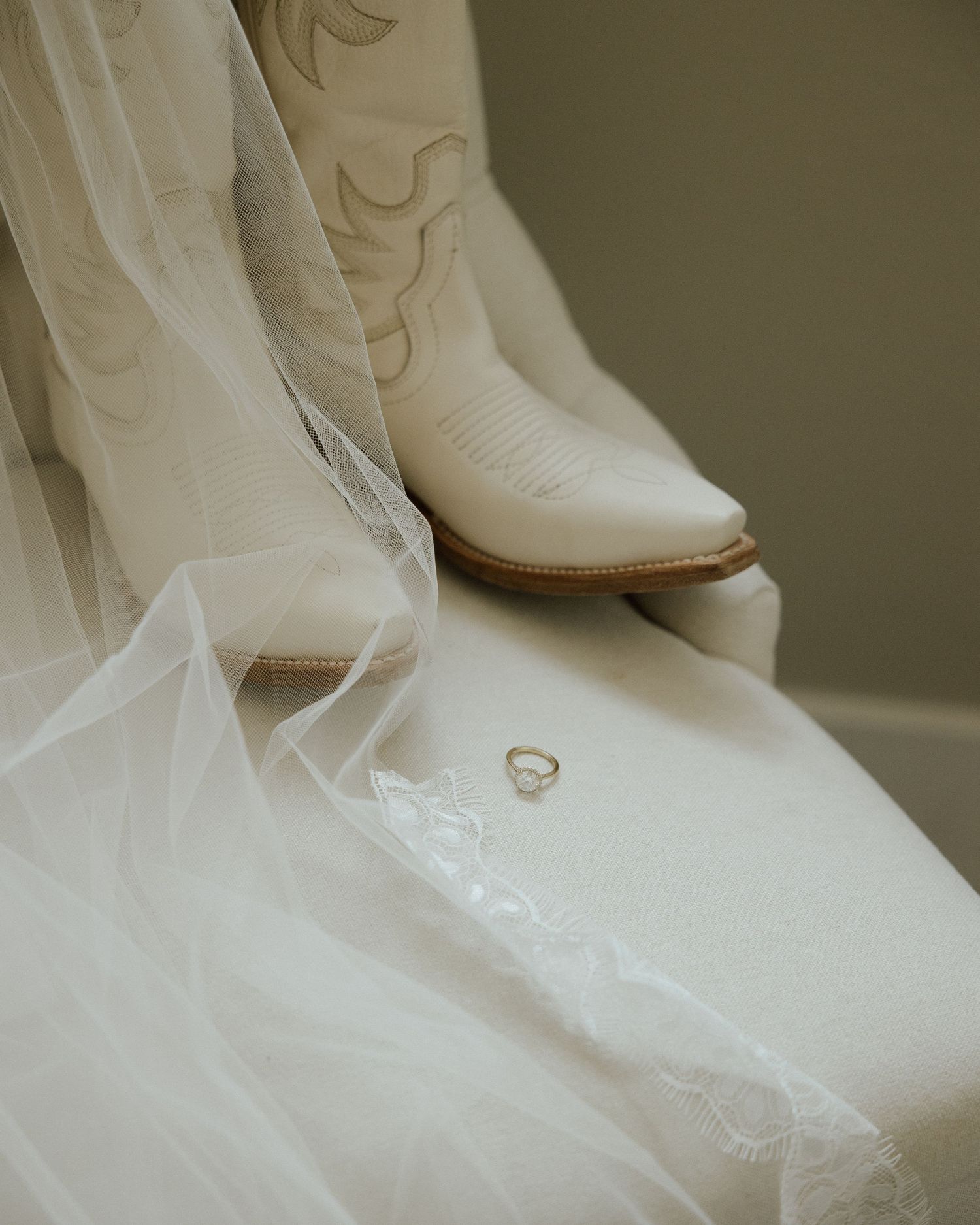 White cowboy boots and wedding veil with wedding ring placed delicately on white fabric.