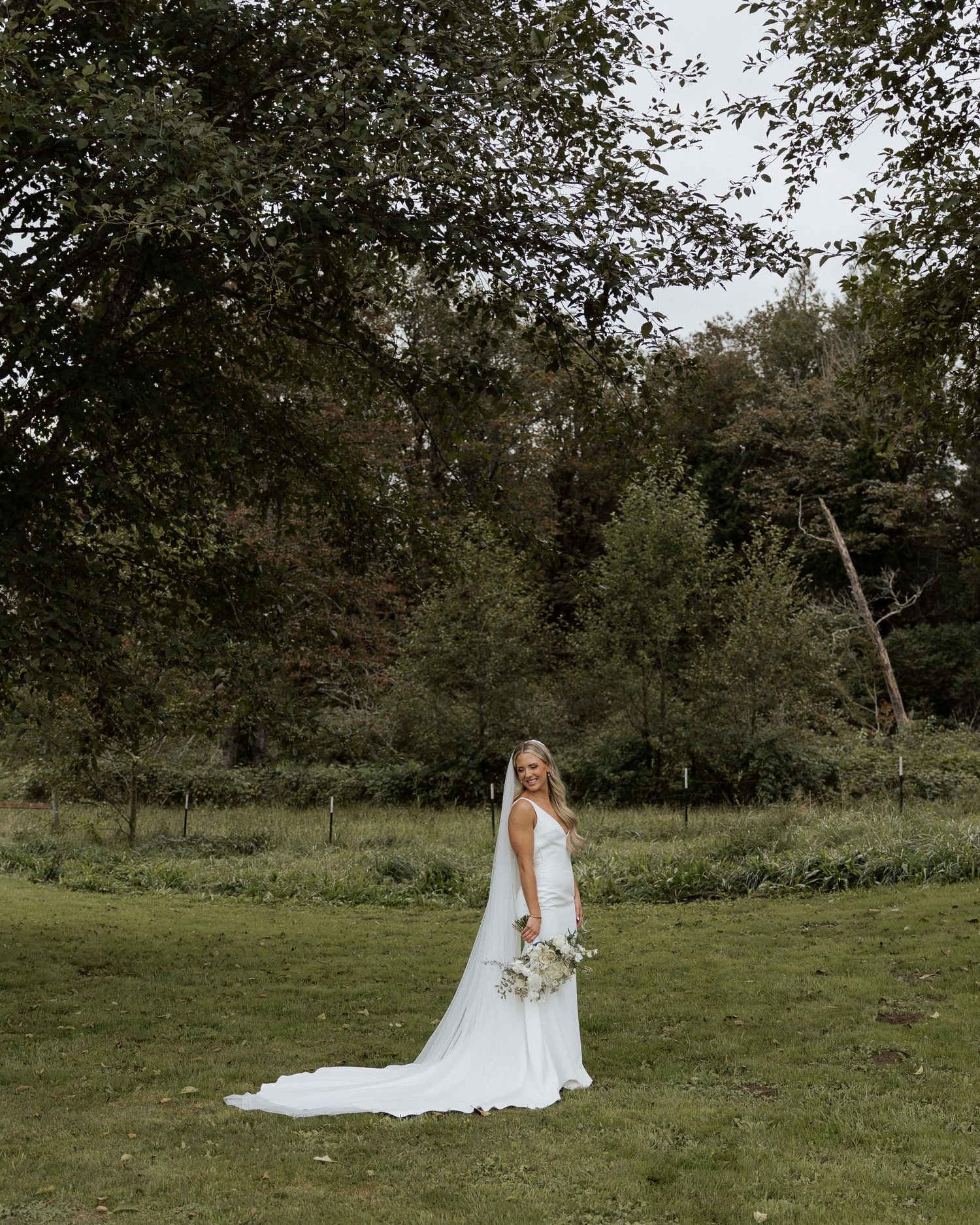 Elegant bride in sleek white gown poses in lush garden setting with trailing veil and white bouquet.