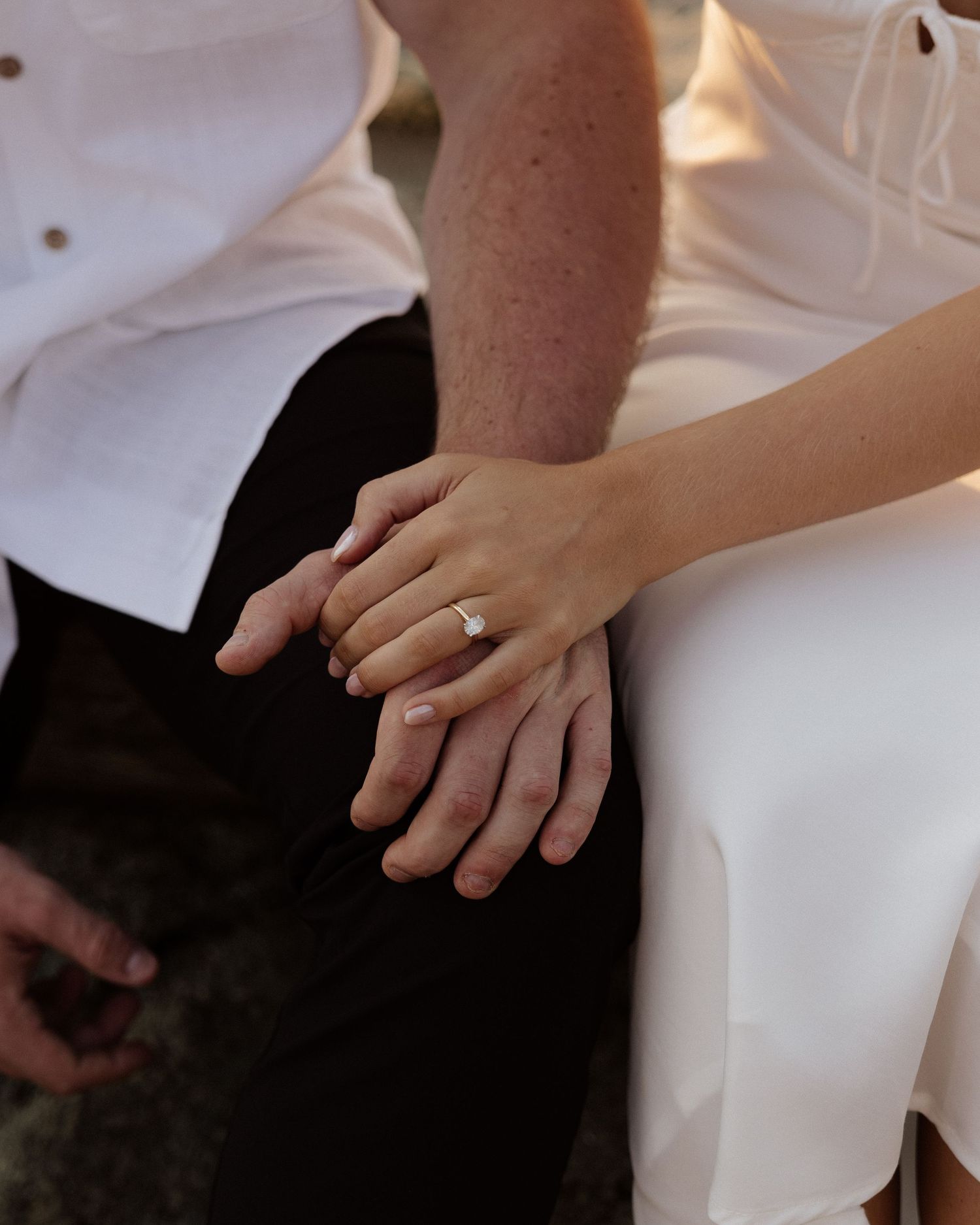 Close-up of hands with engagement ring resting on dark clothing at the beach.