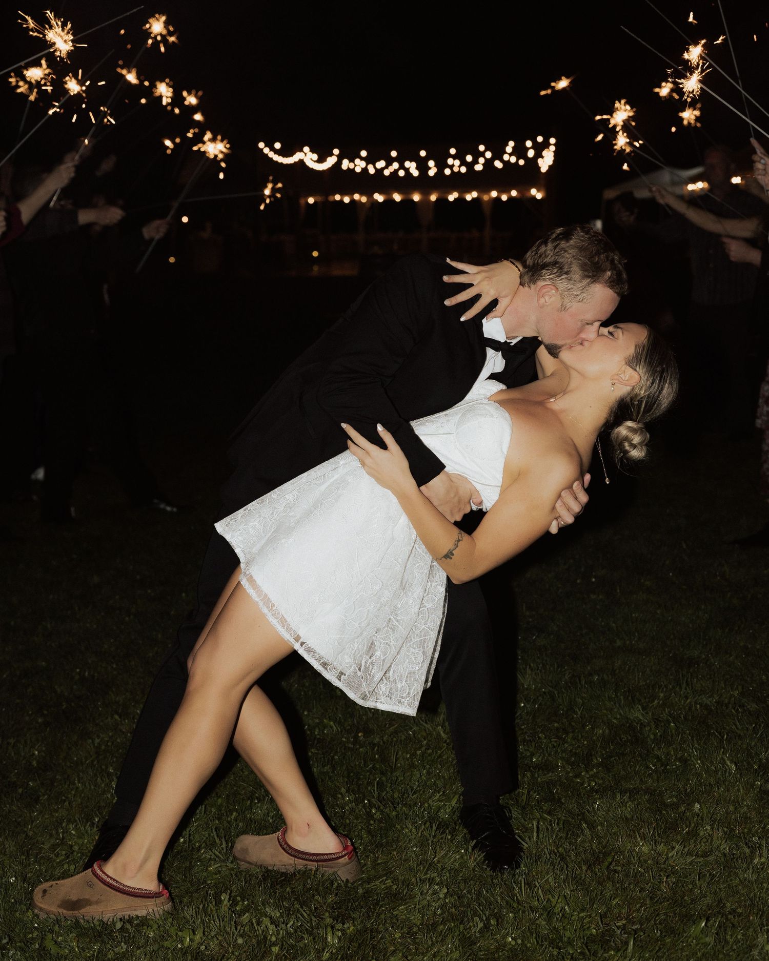 Couple dips and dances under twinkling string lights and sparklers at nighttime celebration.