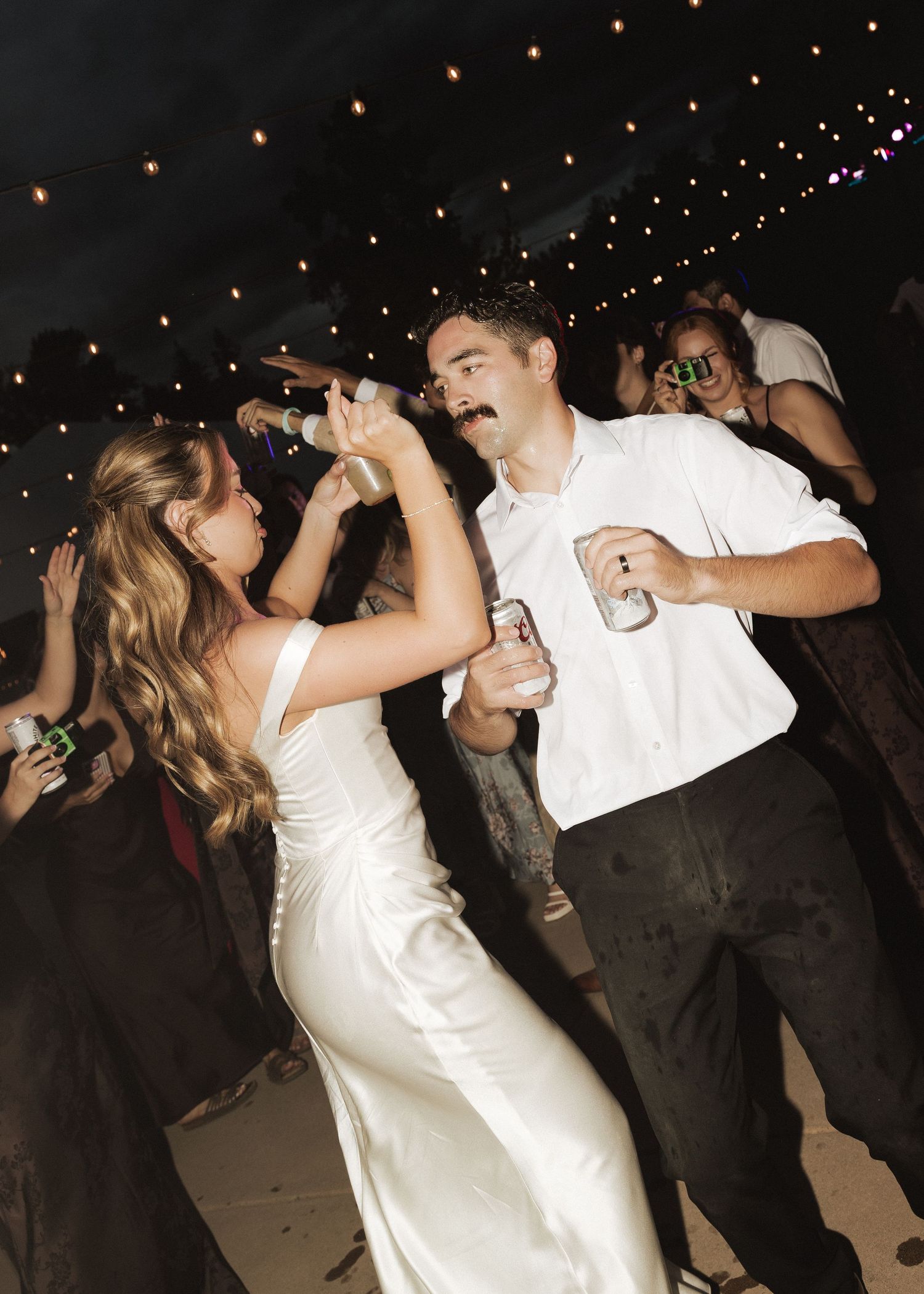 Bride and groom dance joyfully holding drinks at a festive outdoor wedding reception at night.
