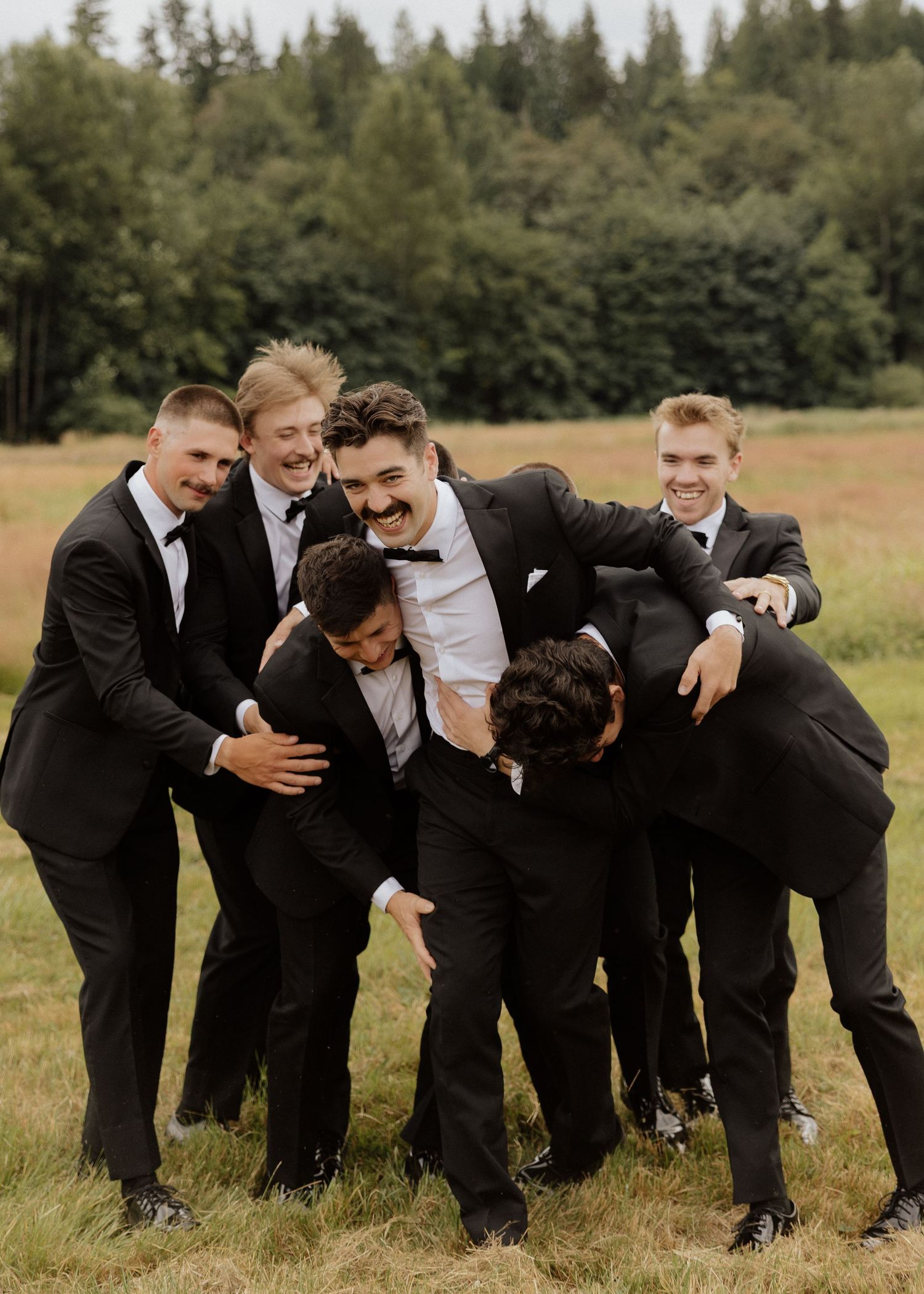 Groomsmen in black suits joyfully huddle around the groom outdoors at a wedding celebration.