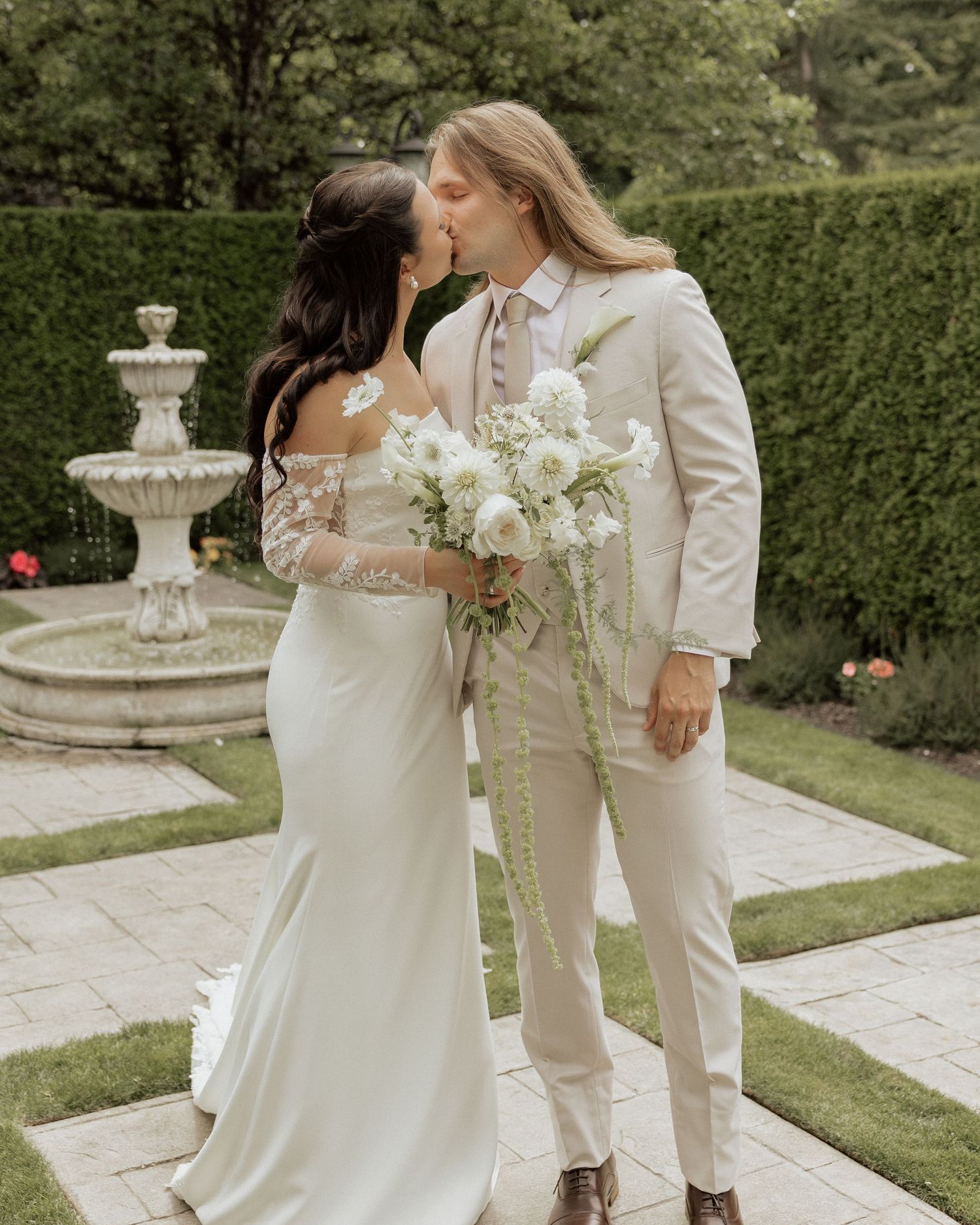 Bride and groom share a kiss in a garden wedding venue, bride holding white floral bouquet, groom in cream suit.