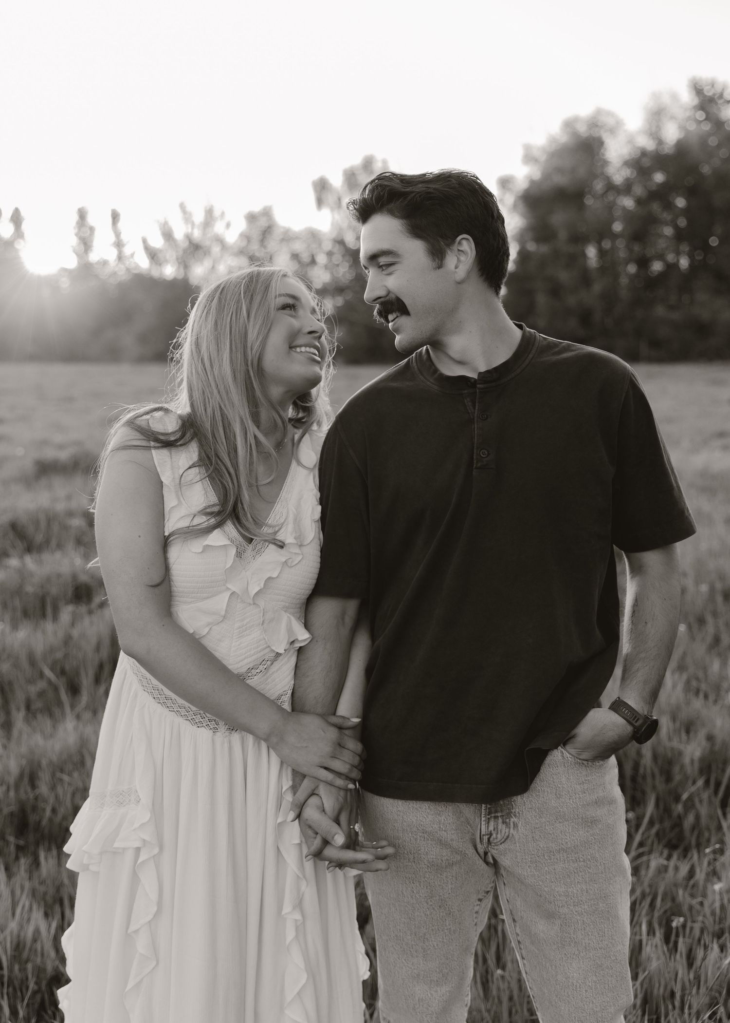 Black and white photo of a smiling couple embracing in a sunlit field, woman in white dress, man in dark shirt.