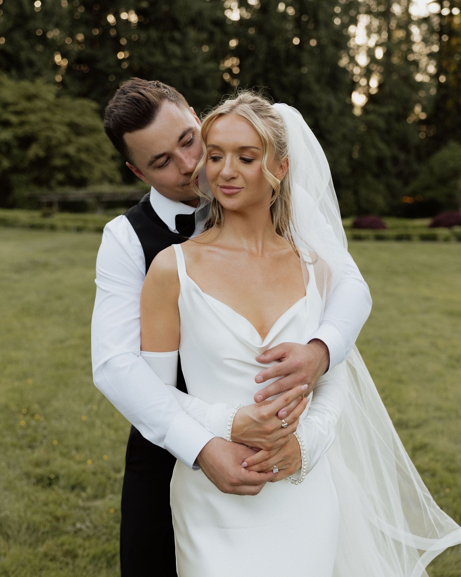 A bride in a white gown and veil embraces her groom in a black tuxedo outdoors at a garden wedding.
