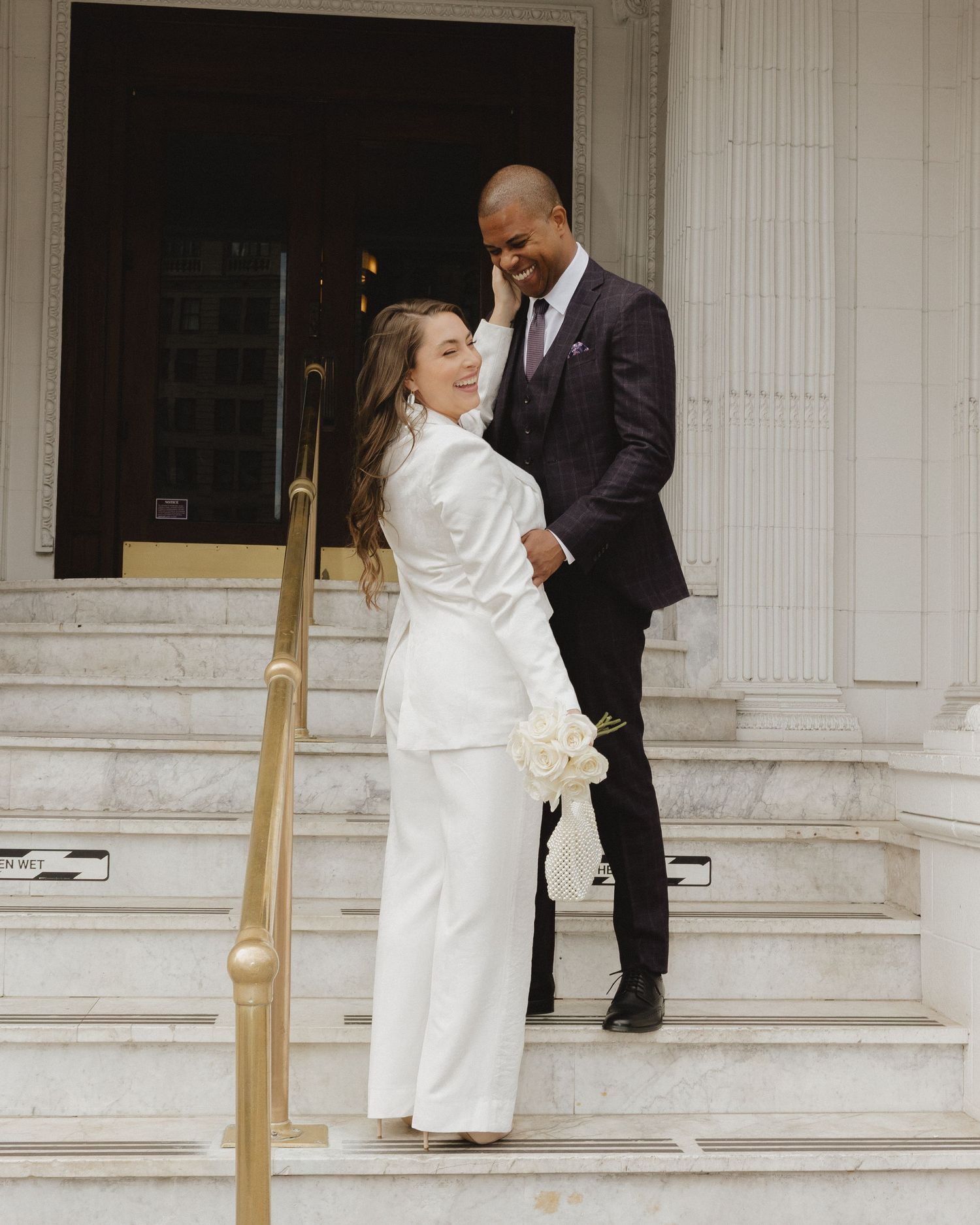 Couple in wedding attire on marble steps, bride in white pantsuit holding bouquet, groom in dark suit smiling.