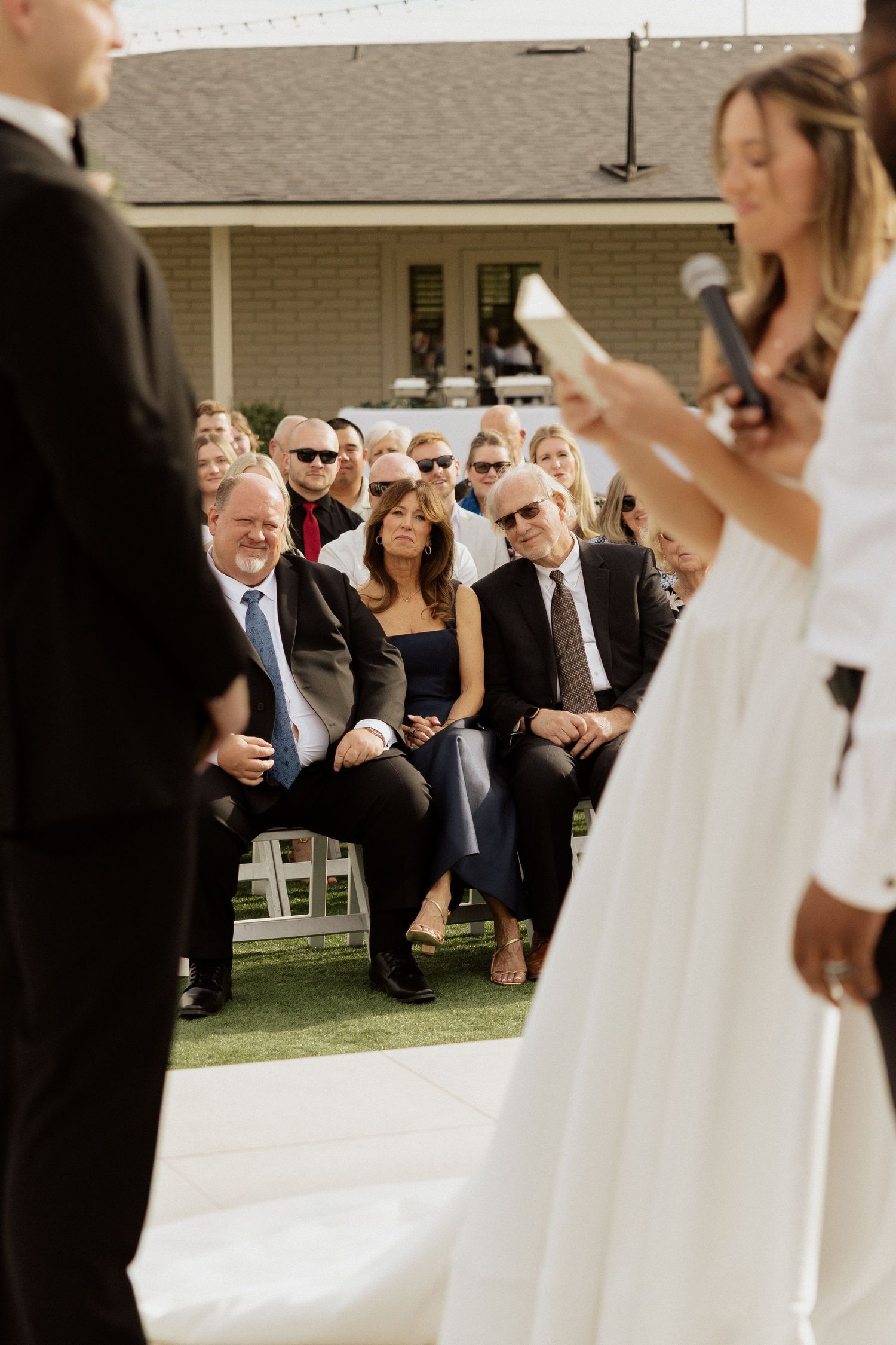 Guests watch attentively as a bride reads vows at an outdoor wedding ceremony on a sunny day.