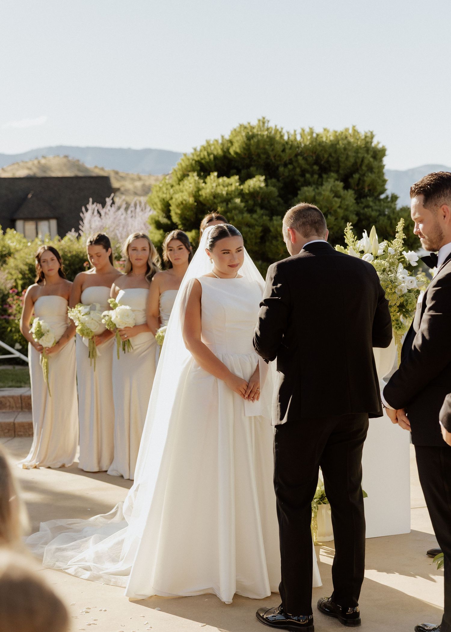 Bride in white gown with veil exchanges vows outdoors, bridesmaids in champagne dresses, mountain backdrop.