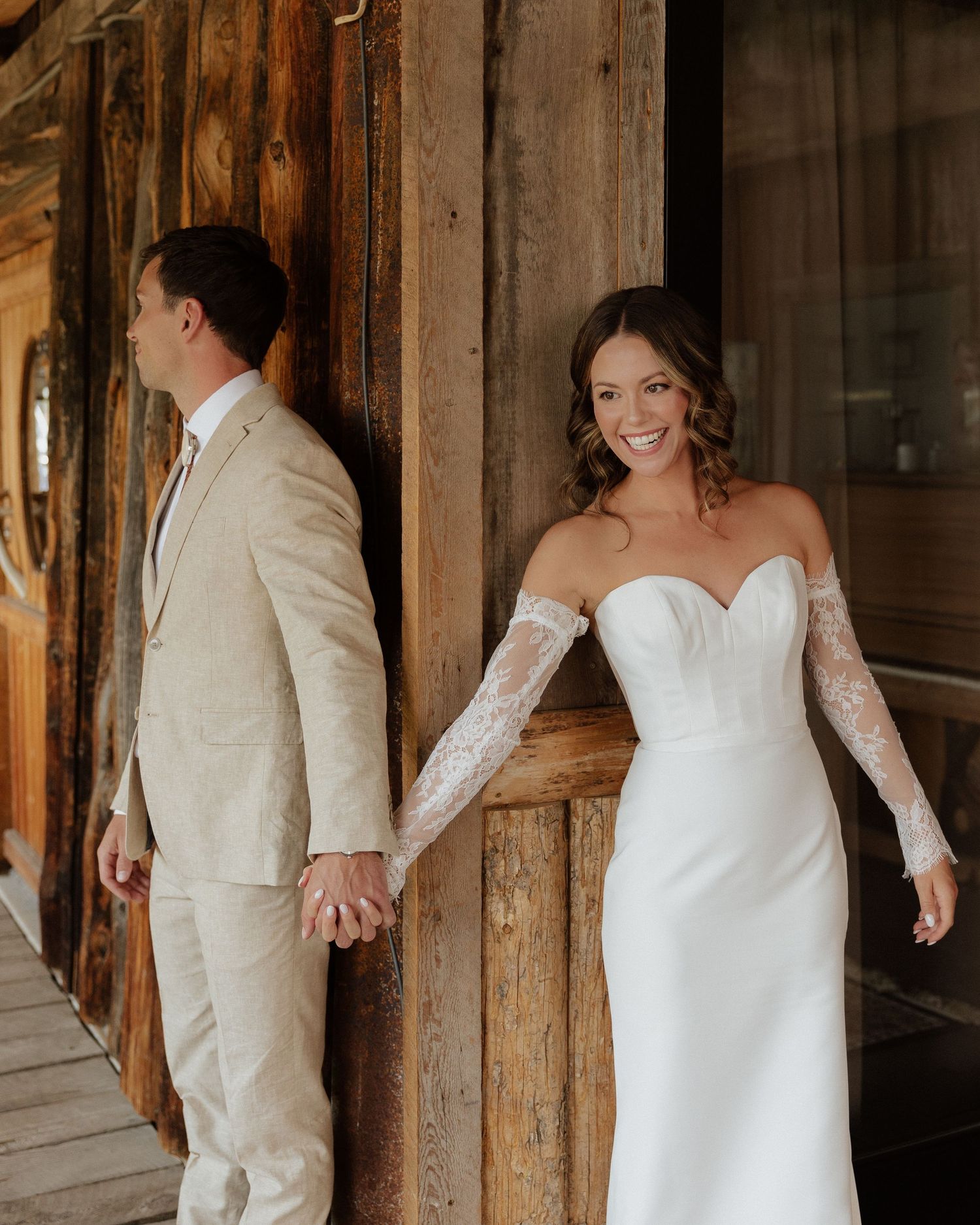 First look wedding photo with couple holding hands while standing on opposite sides of rustic wooden wall.