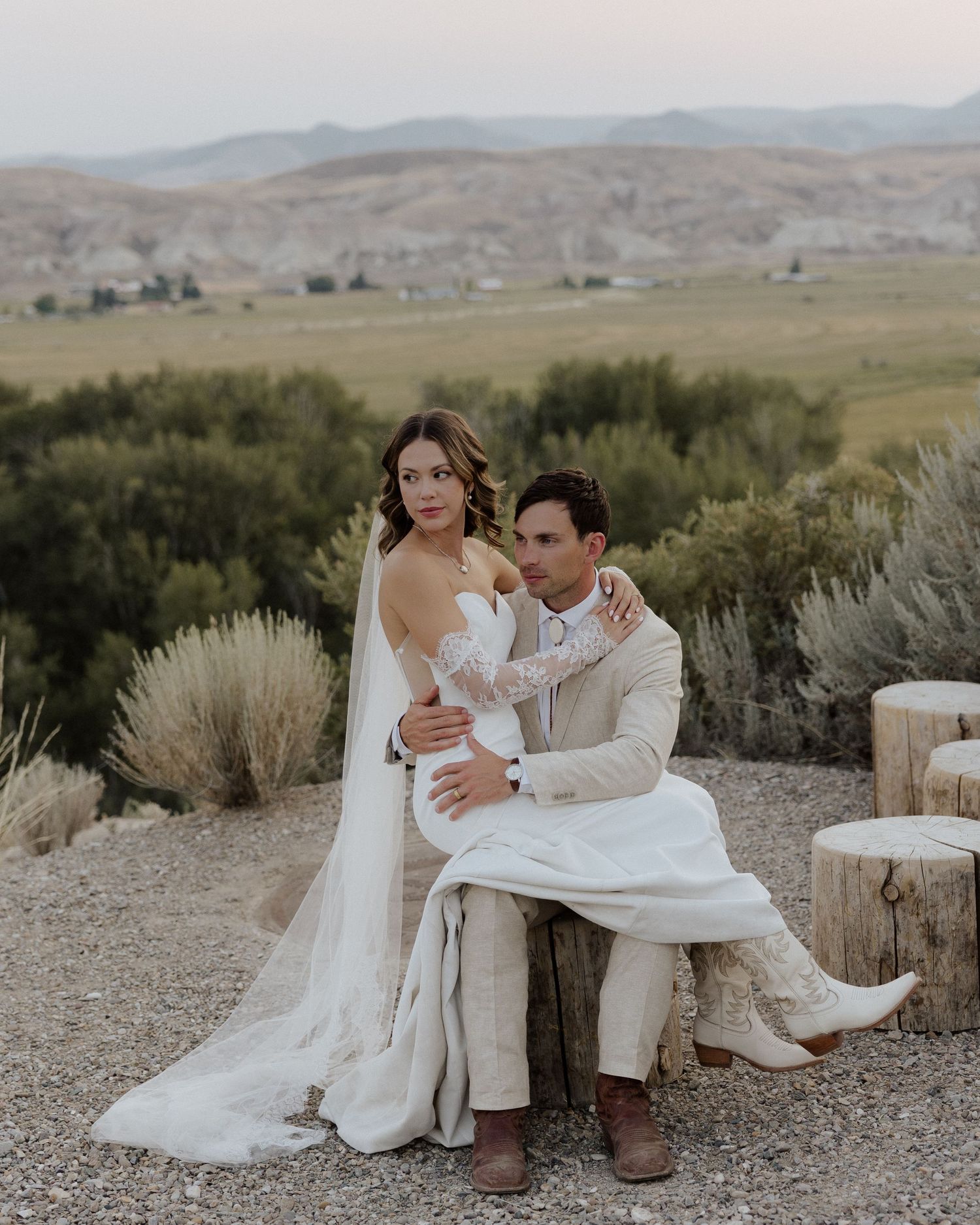 A romantic wedding couple sitting together on a tree stump against a backdrop of rolling hills at sunset.